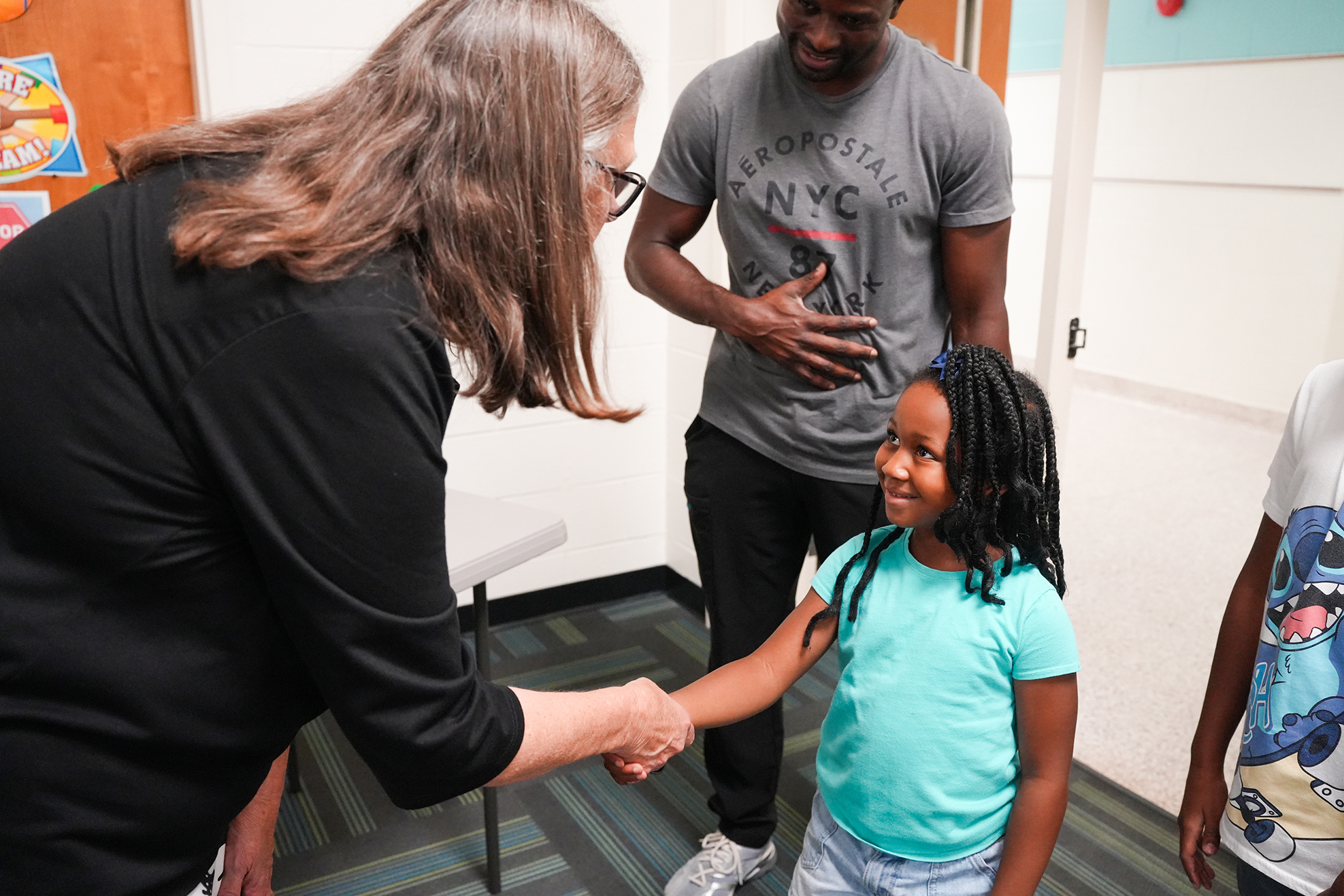 A woman in black shakes hands with a young girl in a light blue shirt. Behind them, a man in gray stands next to a door.