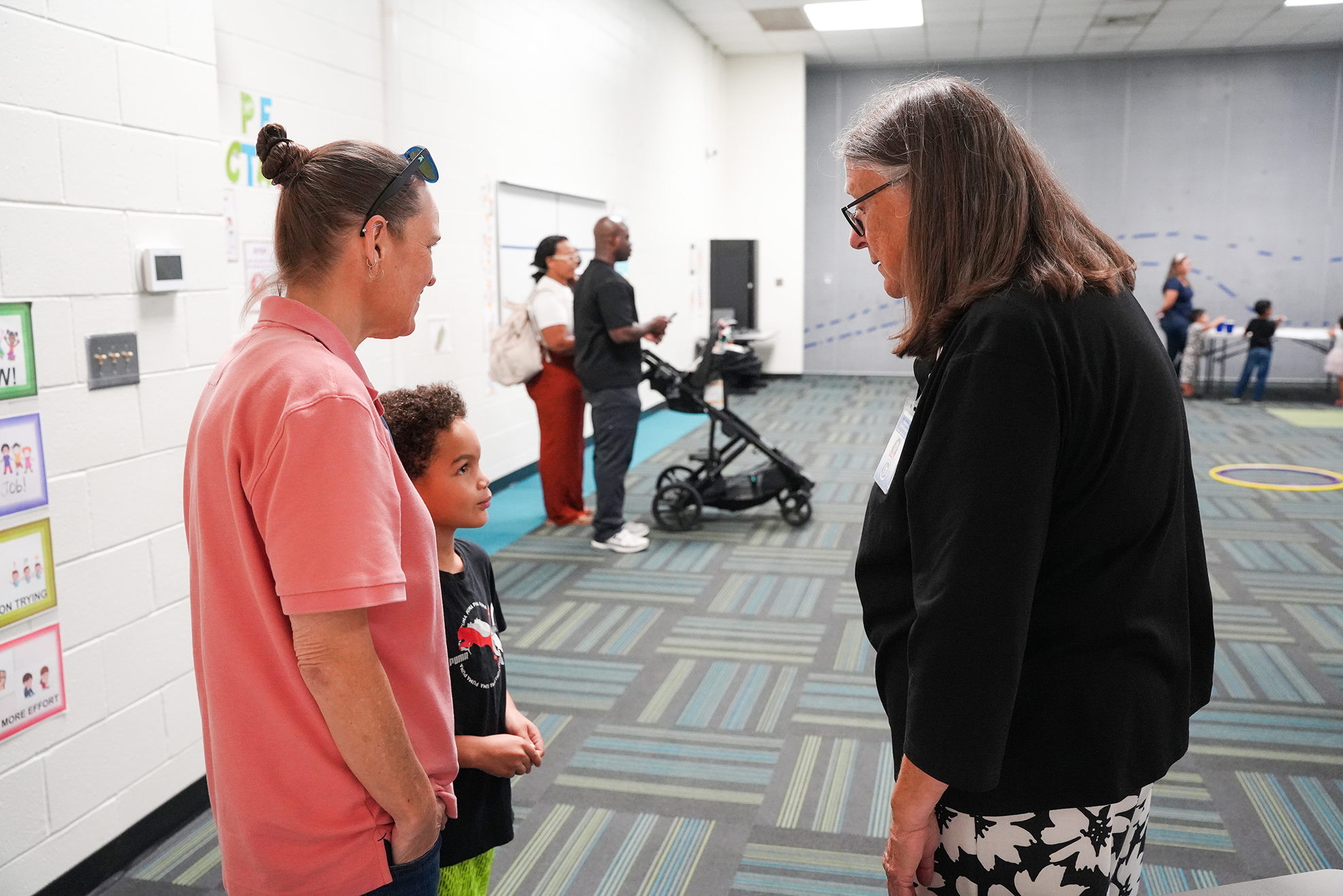 A group of people in a hallway, with one person in a pink shirt and another in black, conversing near a whiteboard.