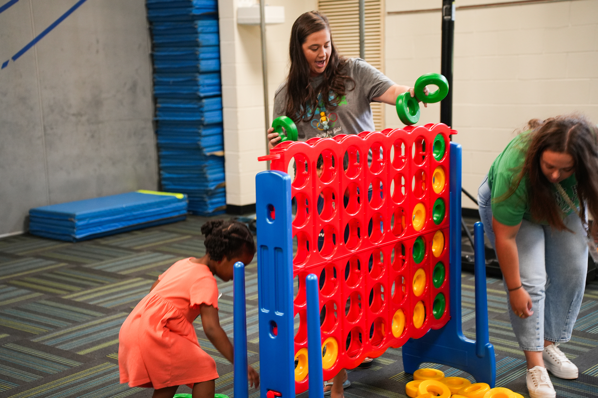 Two adults and a child play a large Connect Four game in a gym, with blue and red equipment.