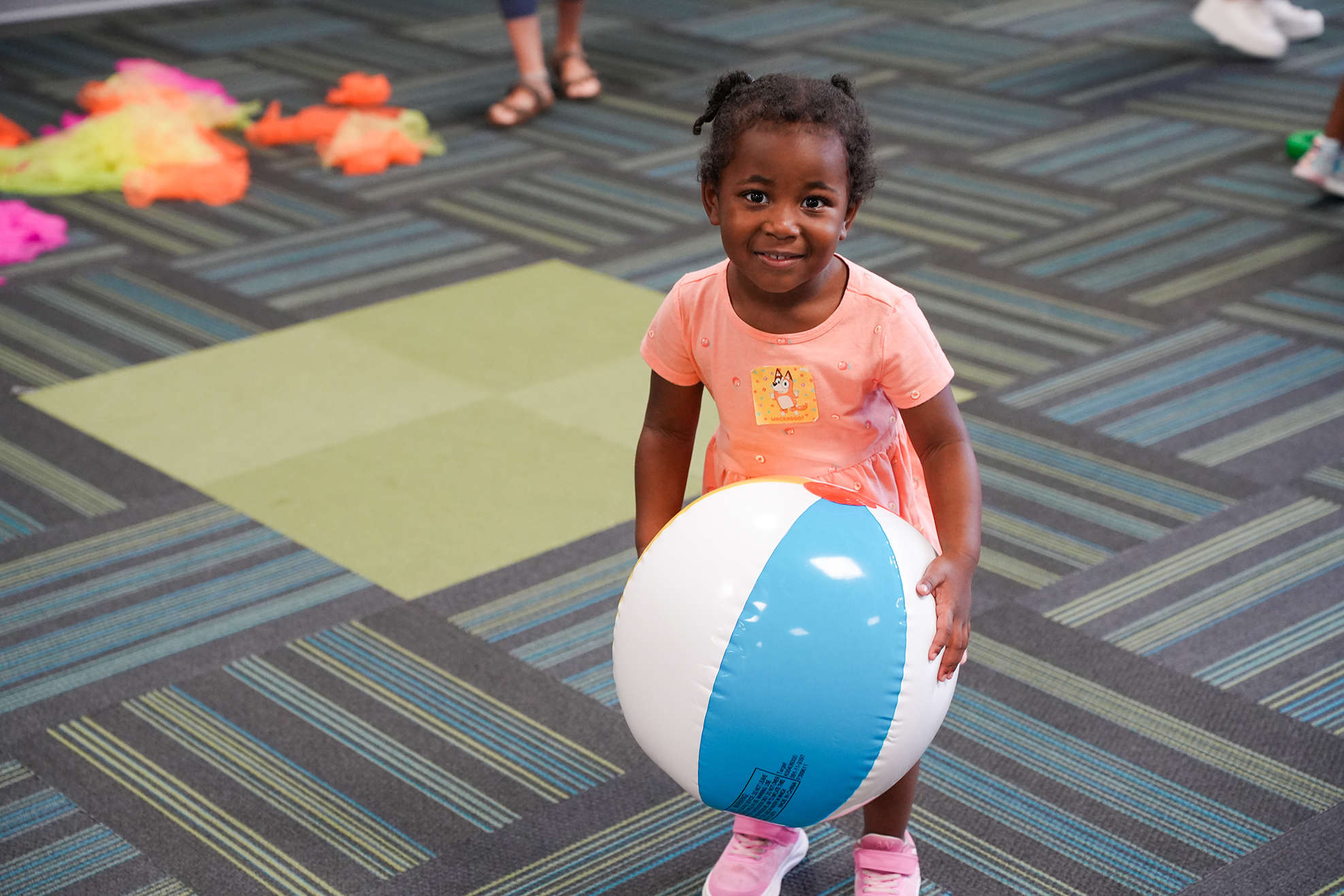 A young child stands on a patterned floor, holding a large beach ball with colorful stripes.