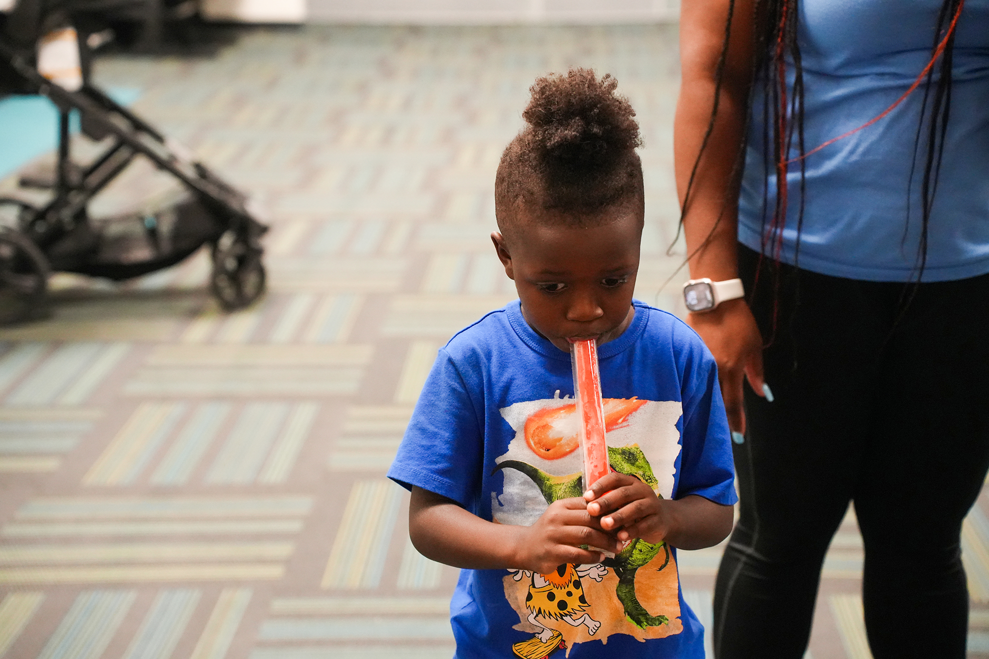 A young child wearing a blue shirt plays a long, thin musical instrument, standing next to an adult.