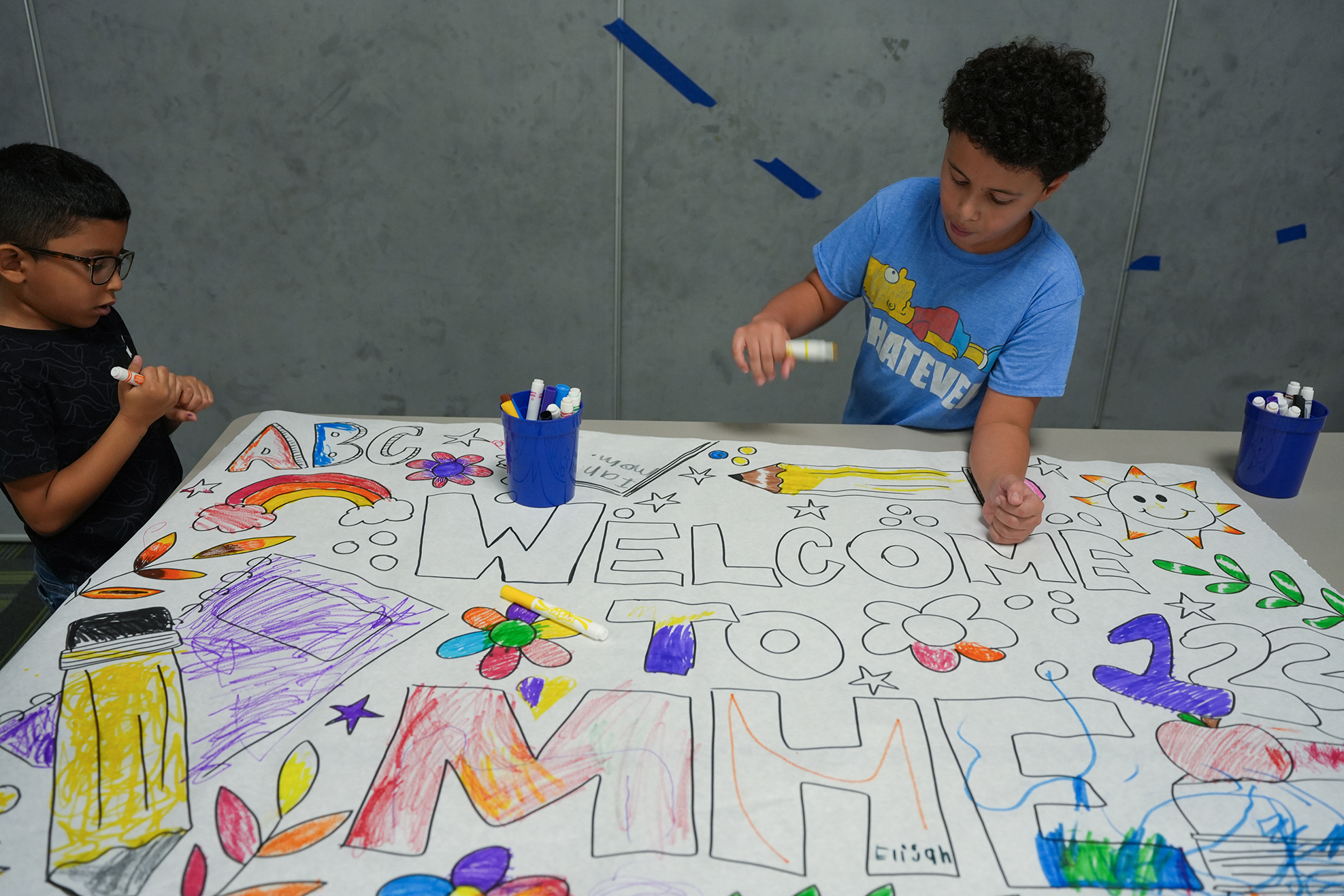 Two kids in a classroom with a large table, creating colorful artwork on a white sheet.