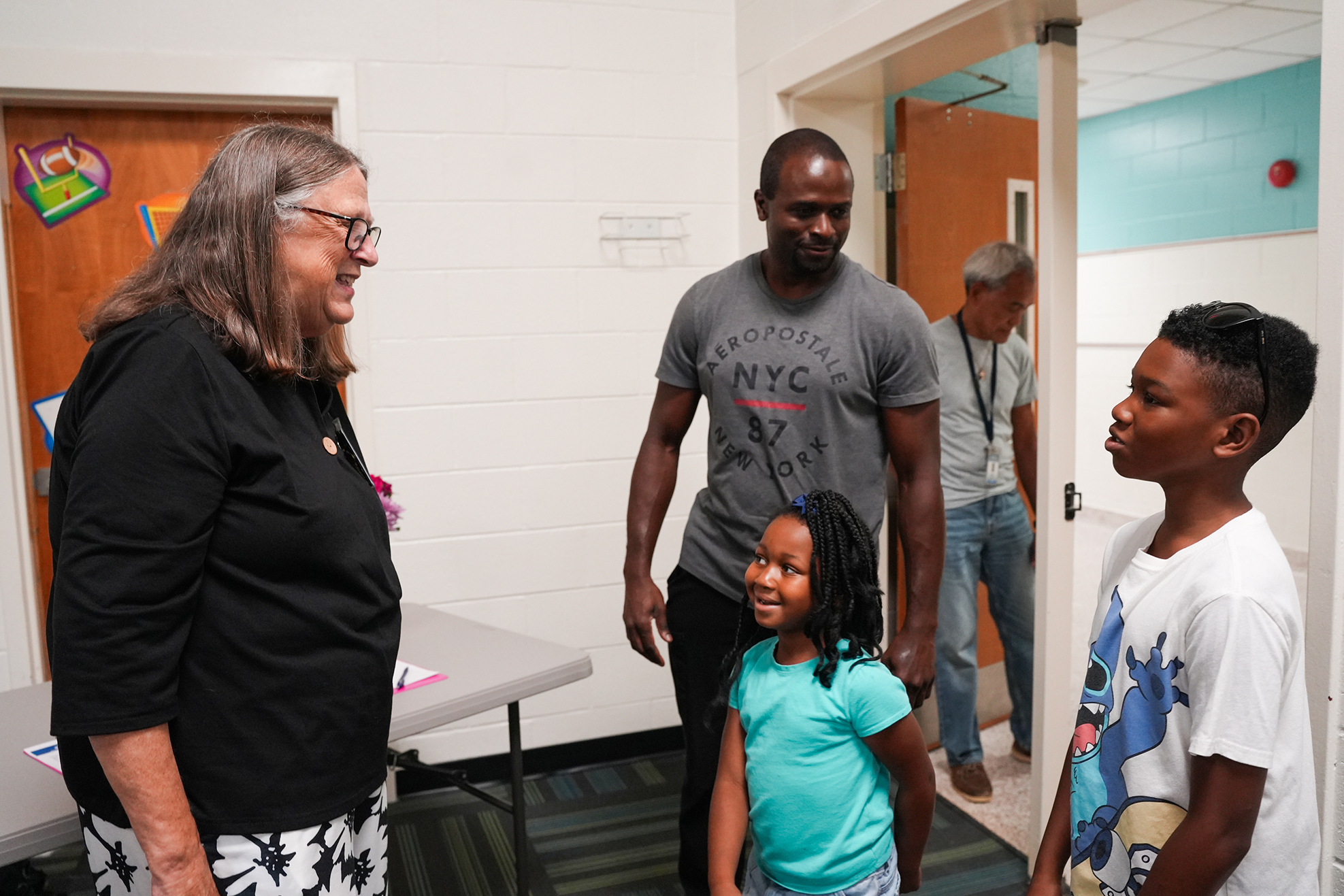 A woman and two children stand in a room with a man, engaged in conversation. Background shows doors and a table.