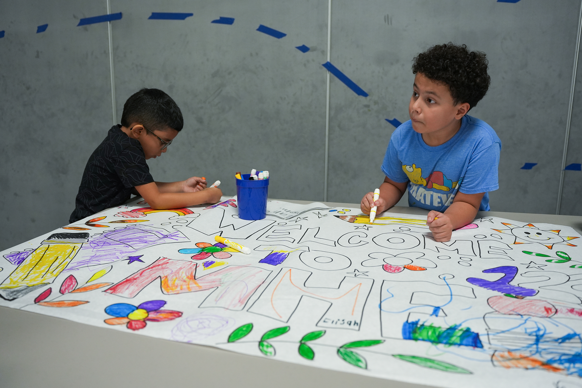 Two children at a table, one wearing glasses, both holding markers. A large, colorful drawing of a "Welcome to the USA" message is on the table.