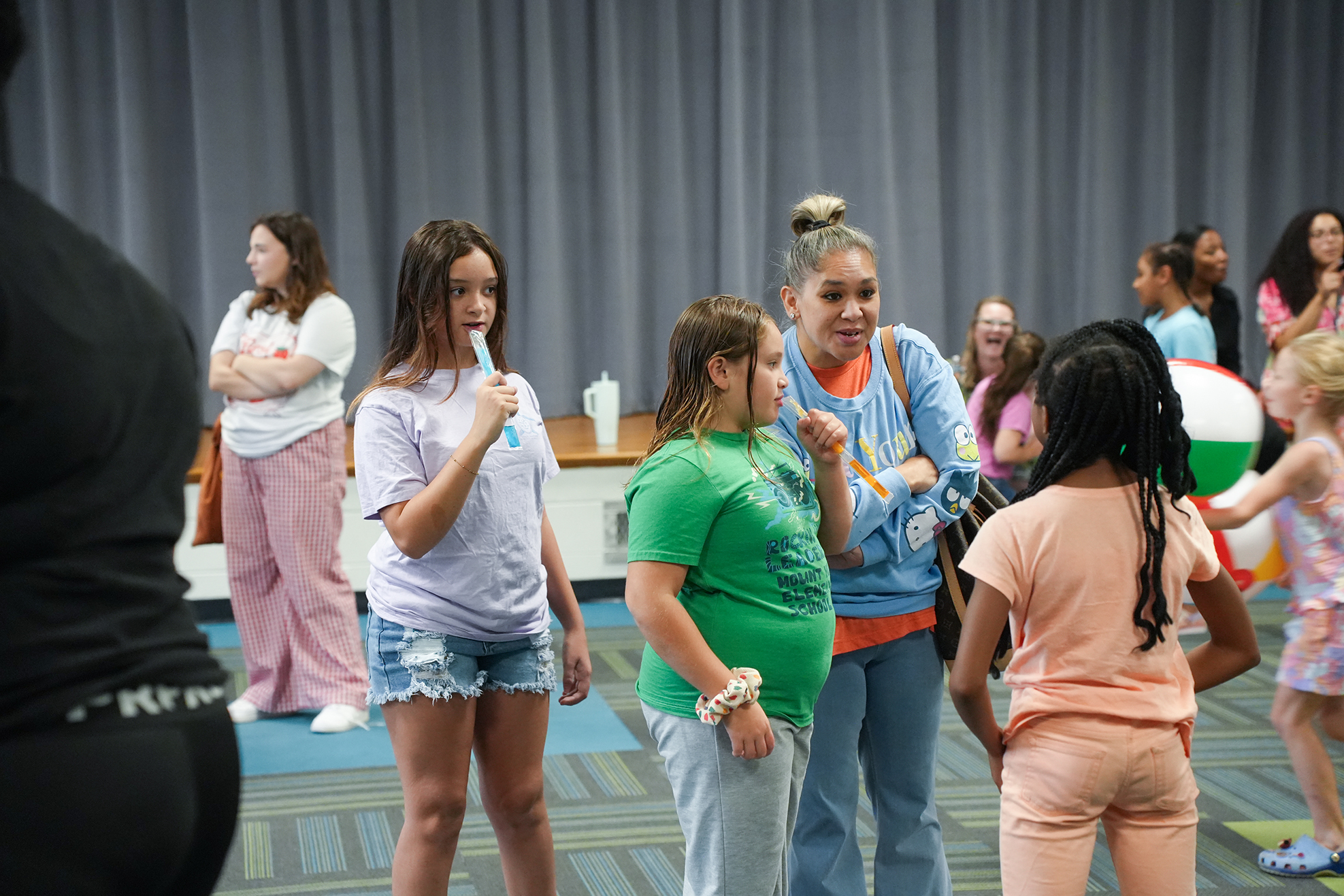 A group of women and girls in a room, some standing in a circle, one holds a toothbrush.