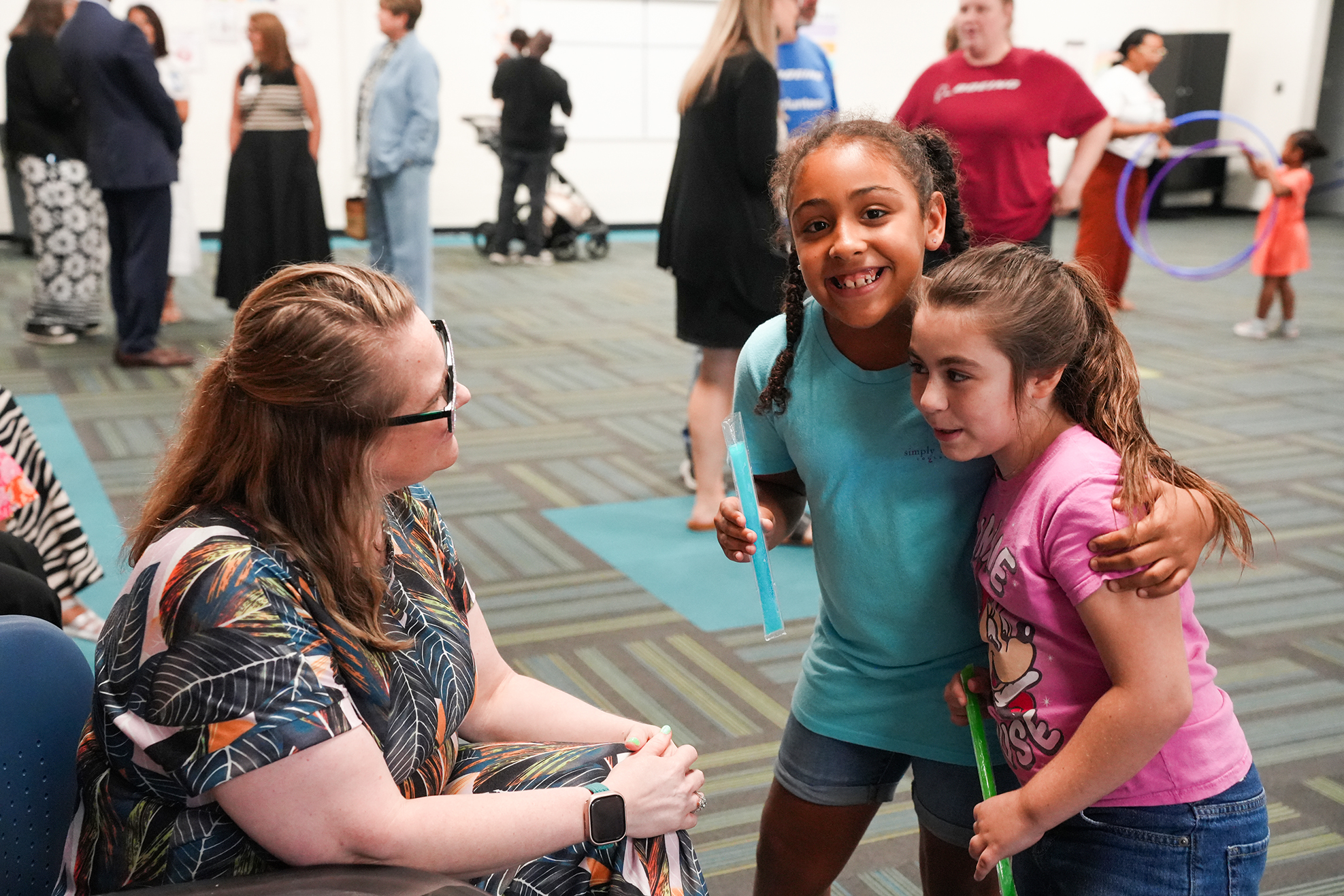 A woman sits with two girls, one holding a hula hoop, while others walk around and interact in a room.