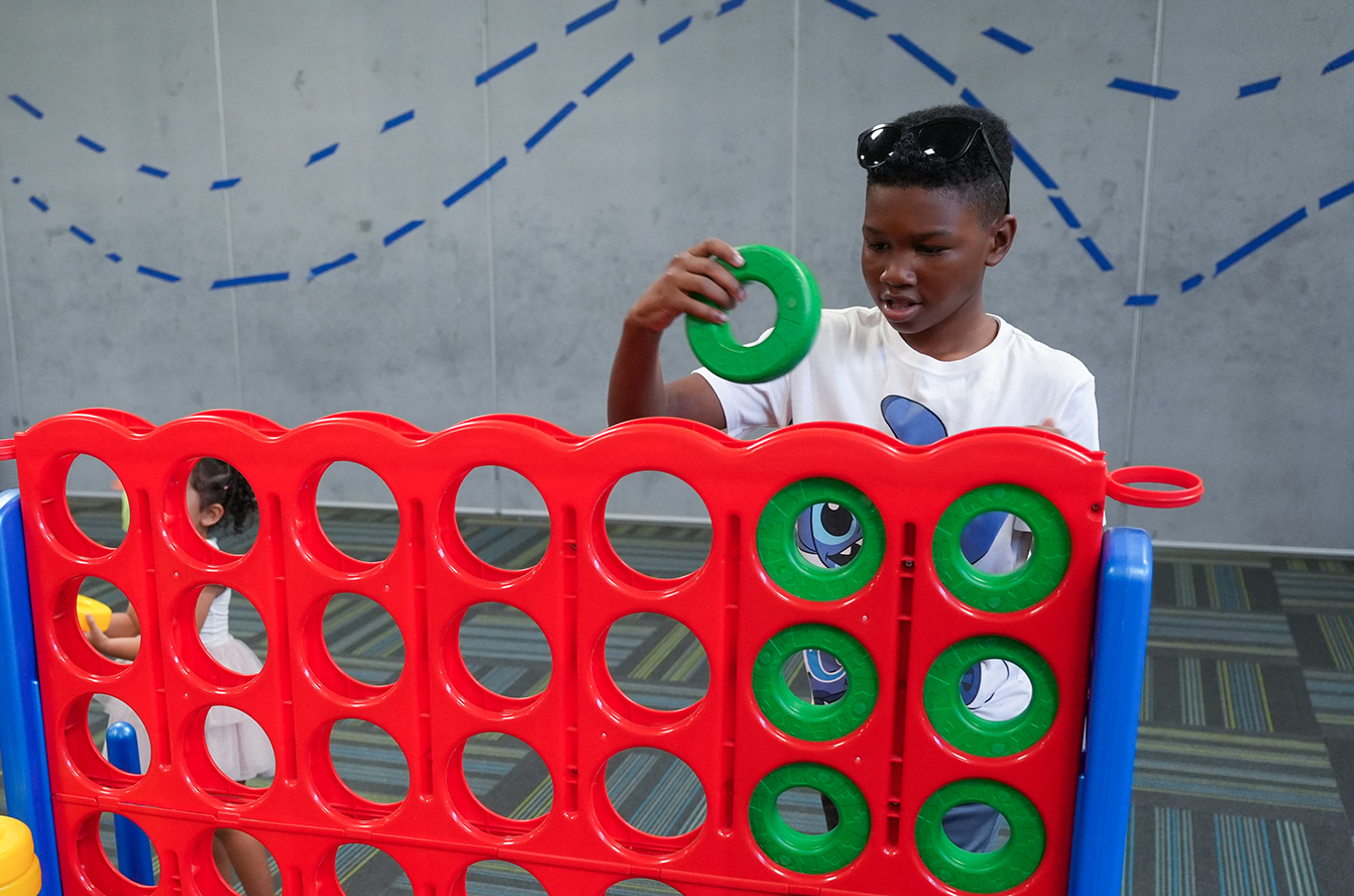 Child holding green ring in front of red, blue, and green Connect Four game in a play area.