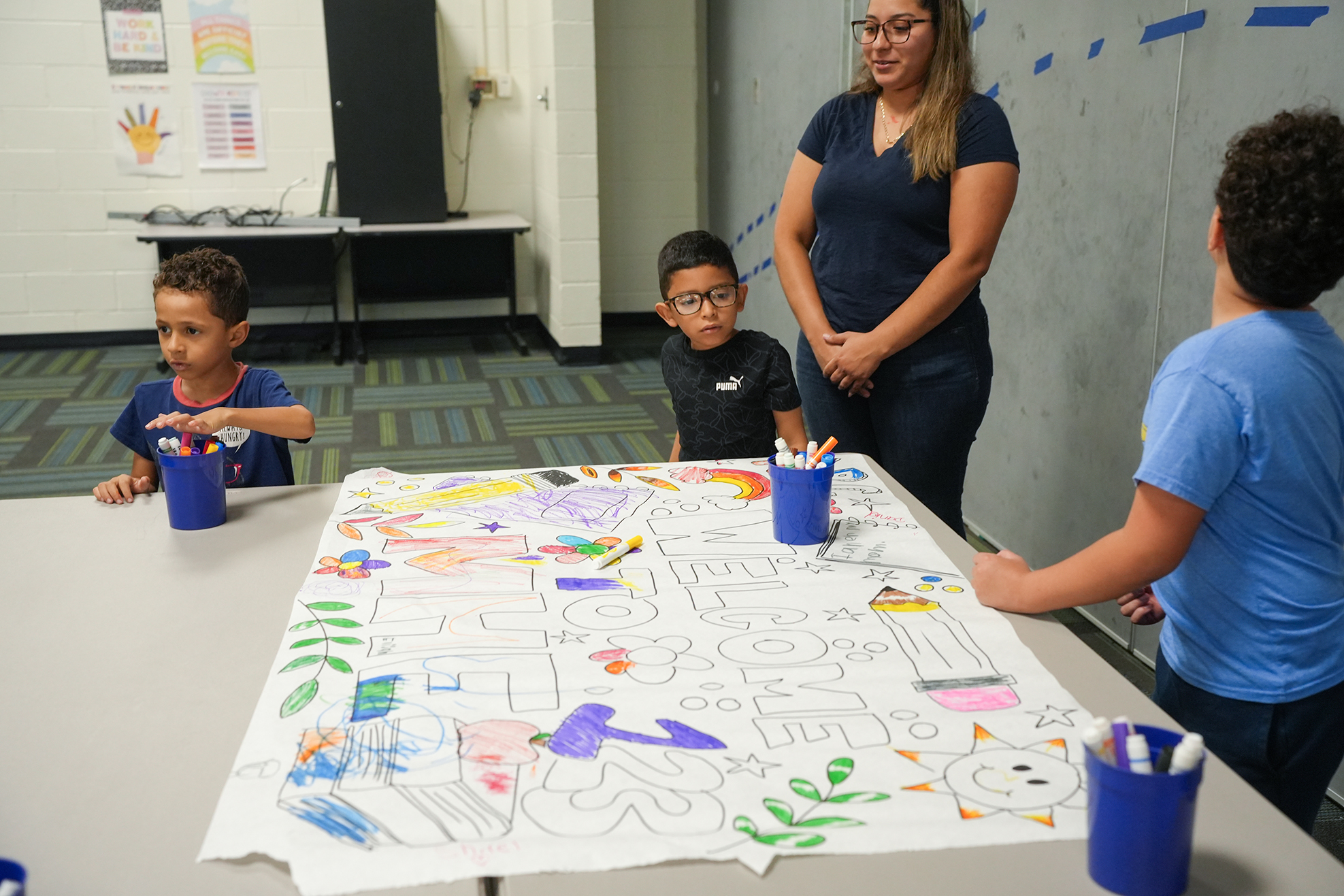 Four children are seated around a table, engaged in coloring a large sheet of paper. One adult stands behind them.