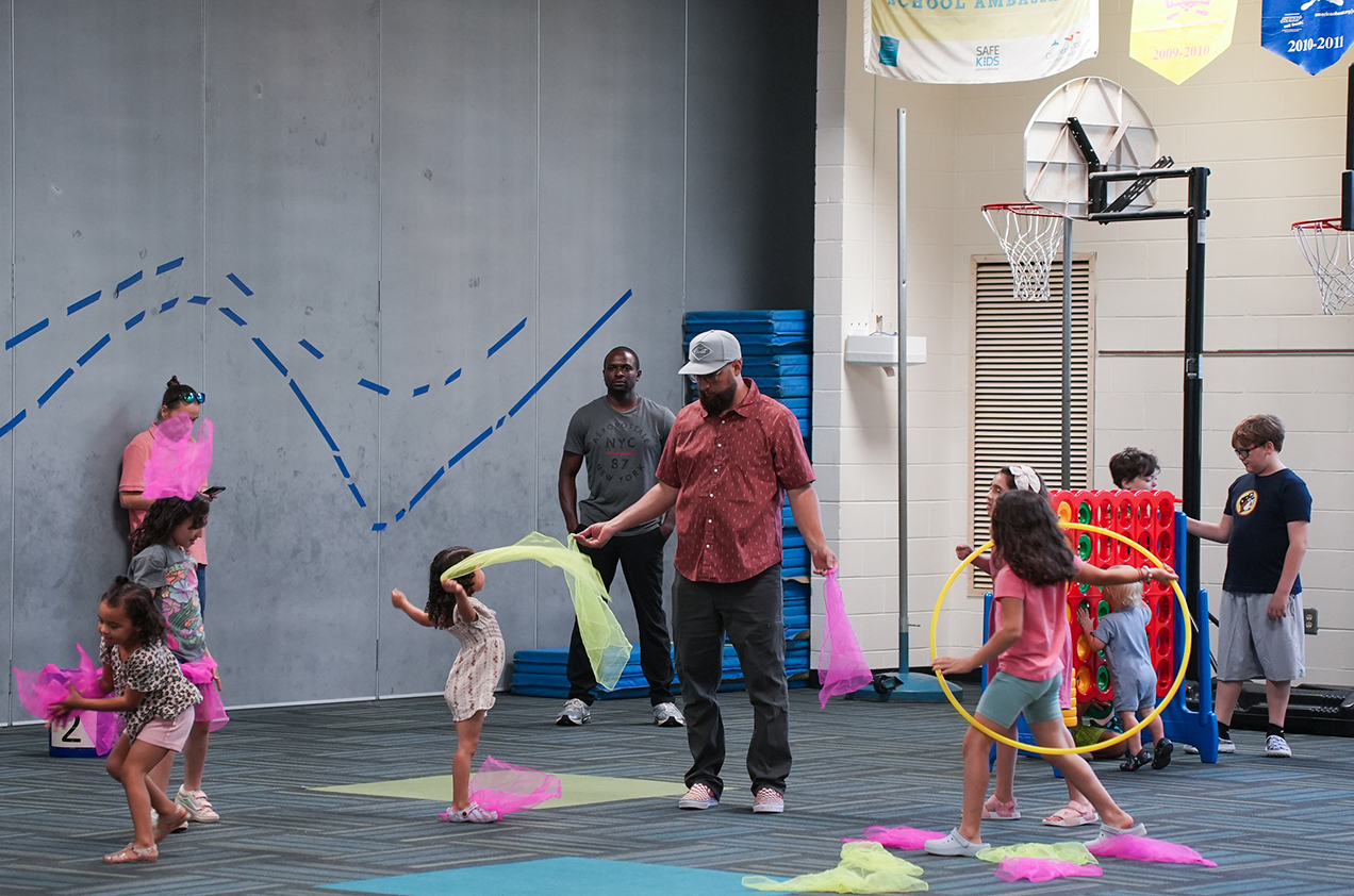 A group of children and adults in a gym, engaging in a dance activity with colorful ribbons and hula hoops.