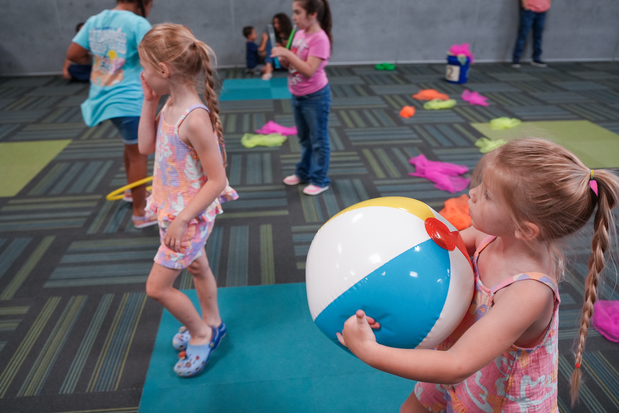 Multiple children play in an indoor area with a ball and other toys. Some wear sandals and shirts.