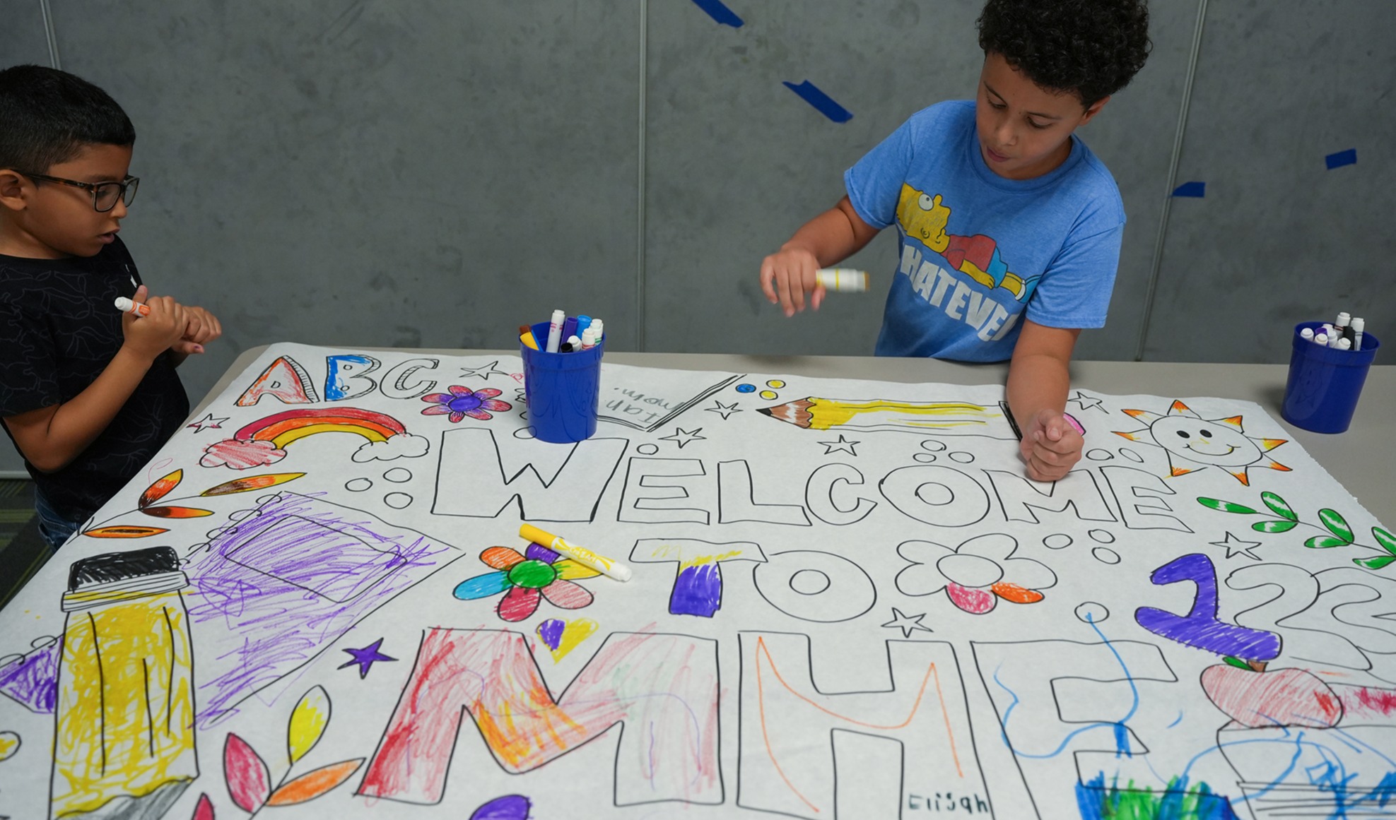 Two children sit at a table with a large white sheet, each using markers to create a colorful design.