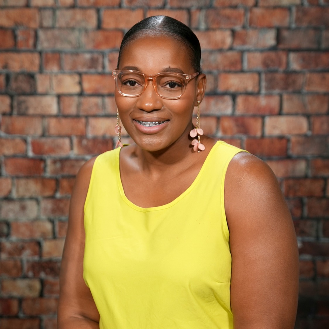 Woman smiling, wearing a yellow sleeveless top, standing in front of a brick wall.
