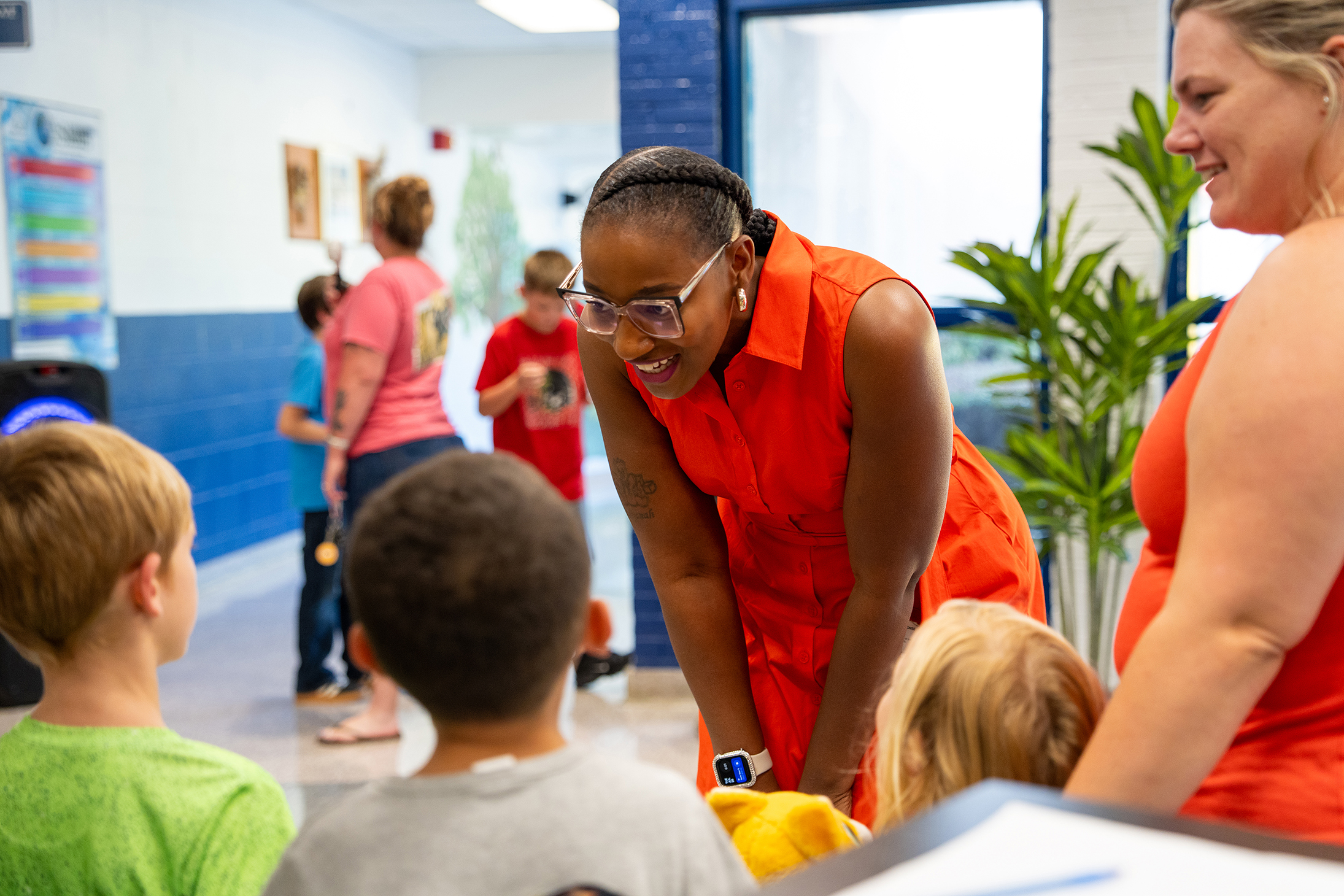Woman in orange dress smiles at children; one wearing green shirt, another in gray, and several others.