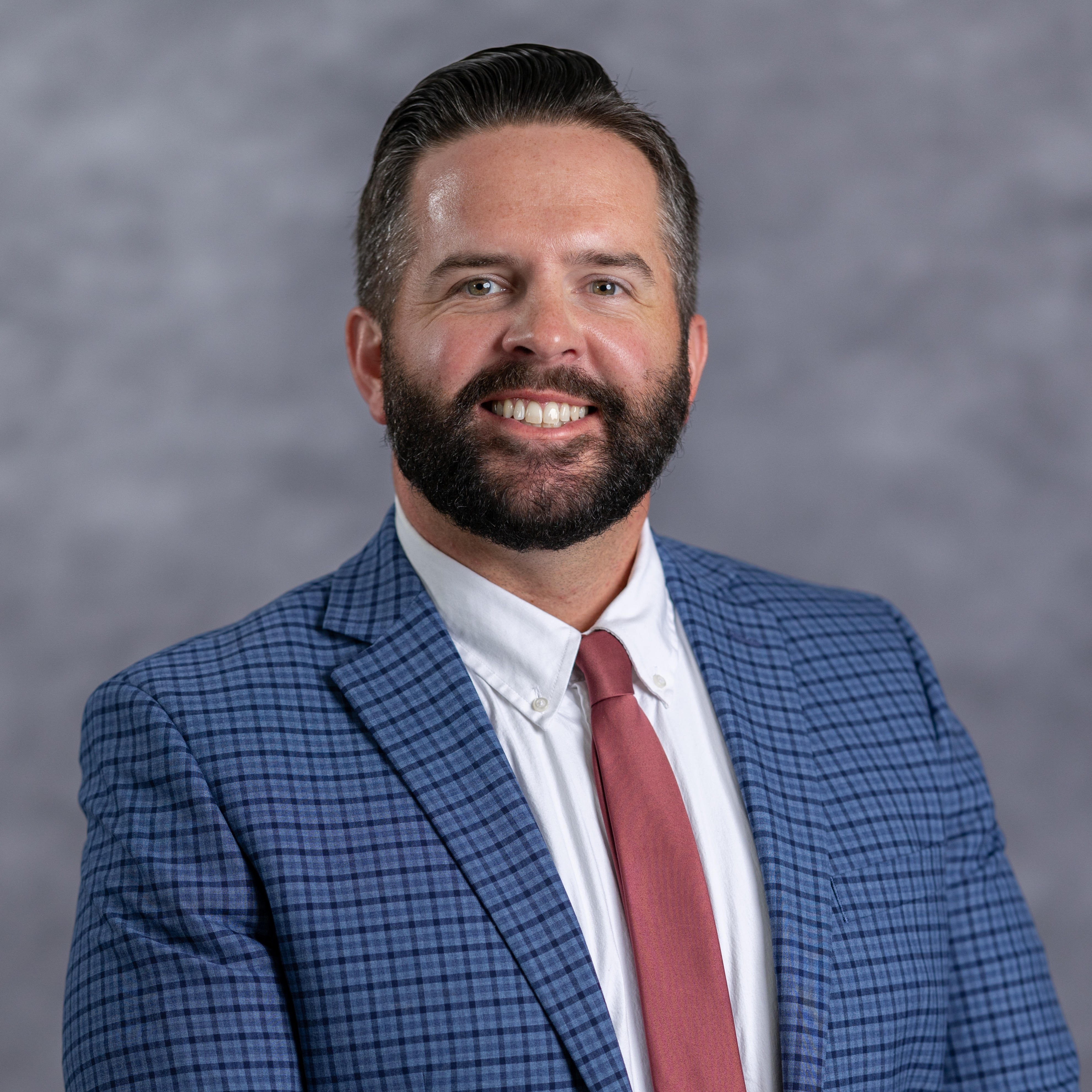 A man with a beard and short hair is smiling. He wears a blue suit with a red tie. The background is gray.