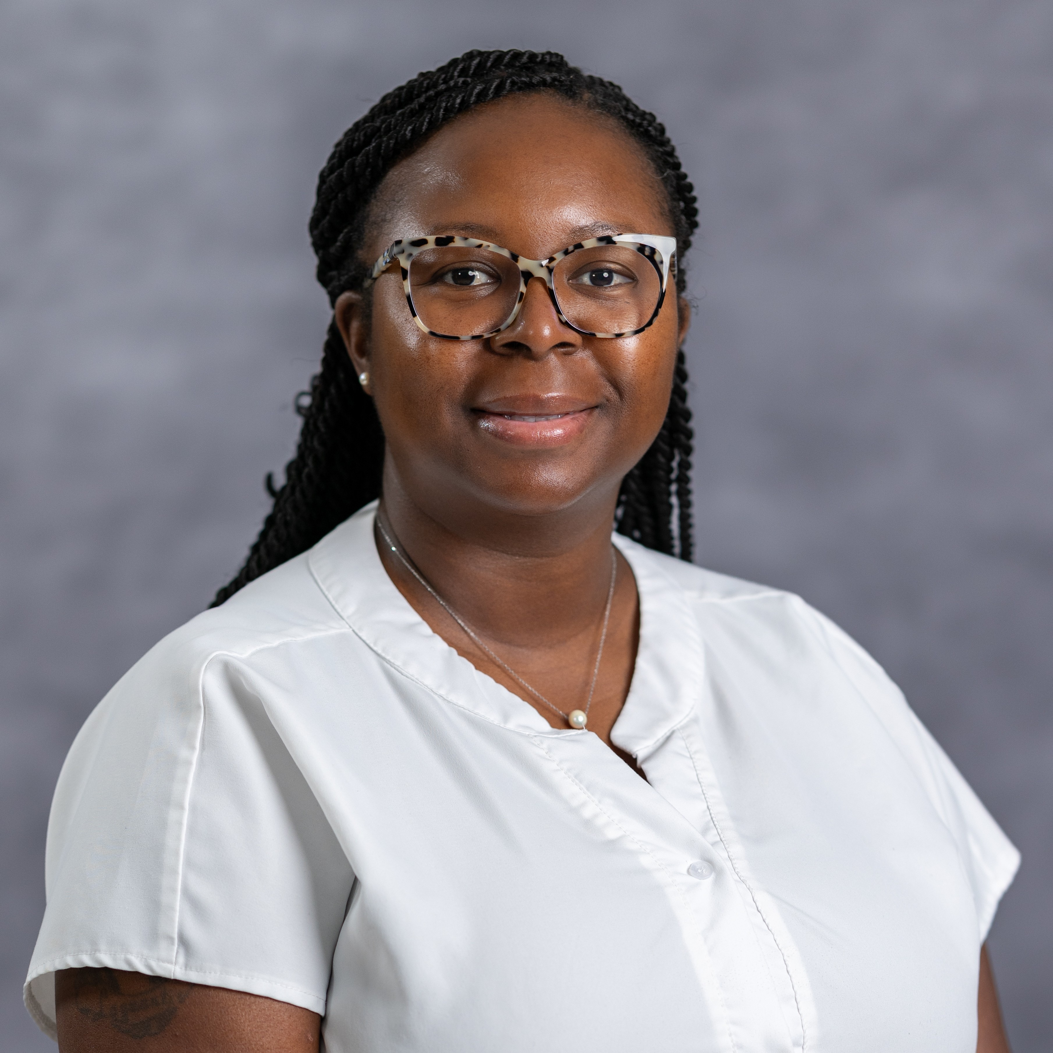 A woman with braided hair and glasses smiles for the camera. She wears a white blouse and a necklace.