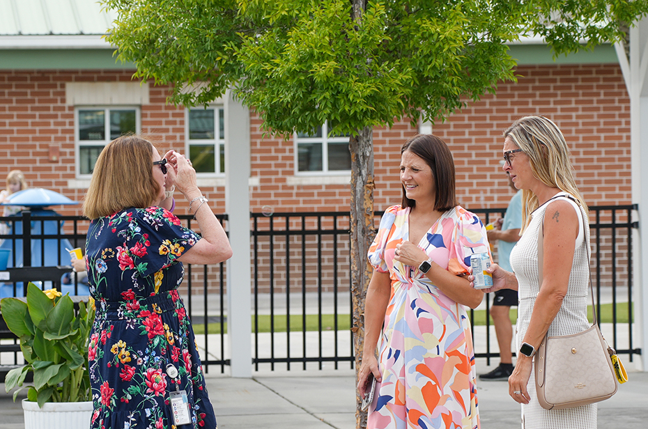Three women standing in front of a brick building with a fence; one takes a photo, one looks at her, and one carries a bag.