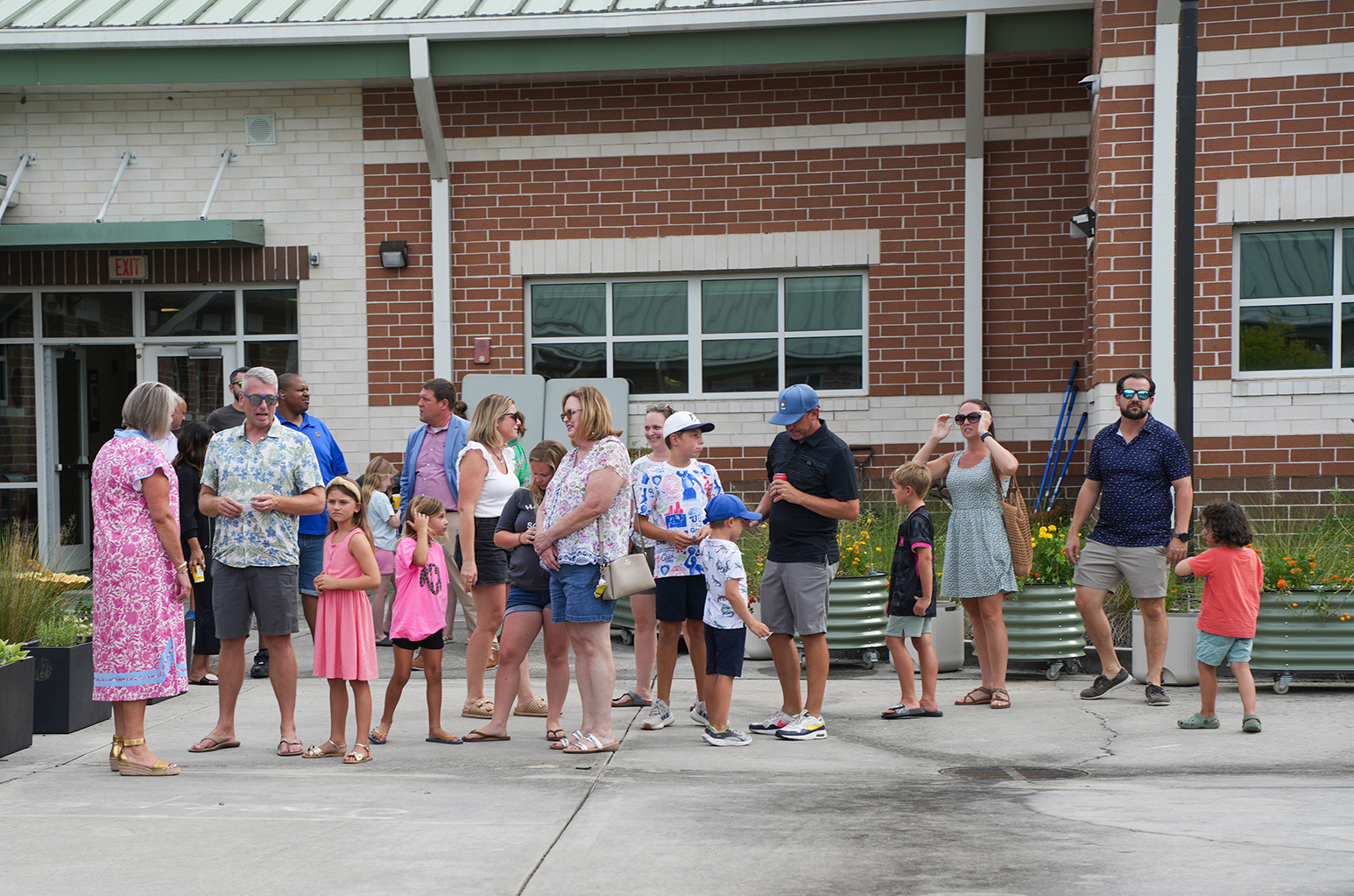 A group of people, some wearing hats, glasses, and sandals, stand in a line outside a brick building.