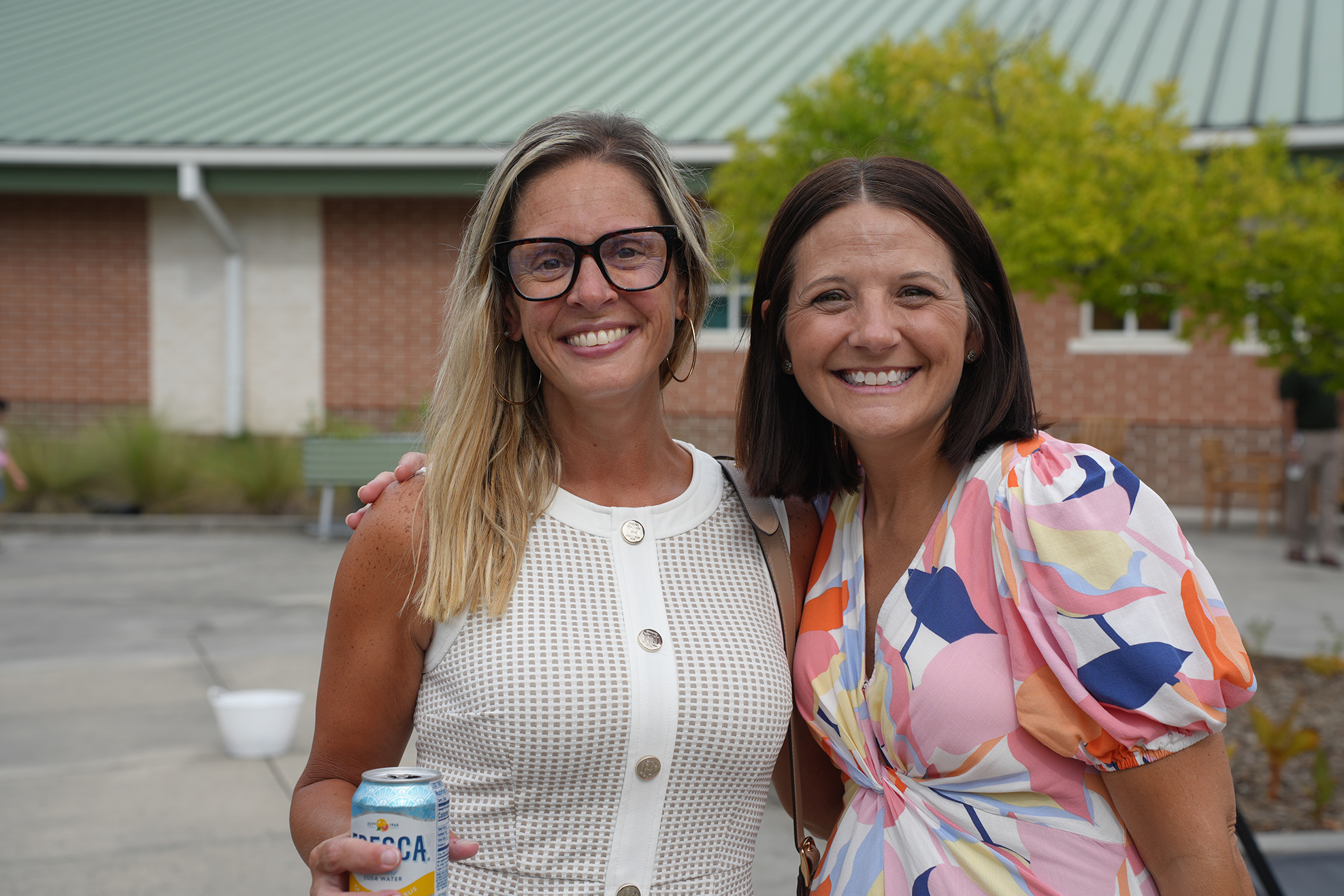 Two smiling women stand together outside near a building. One holds a beverage can.