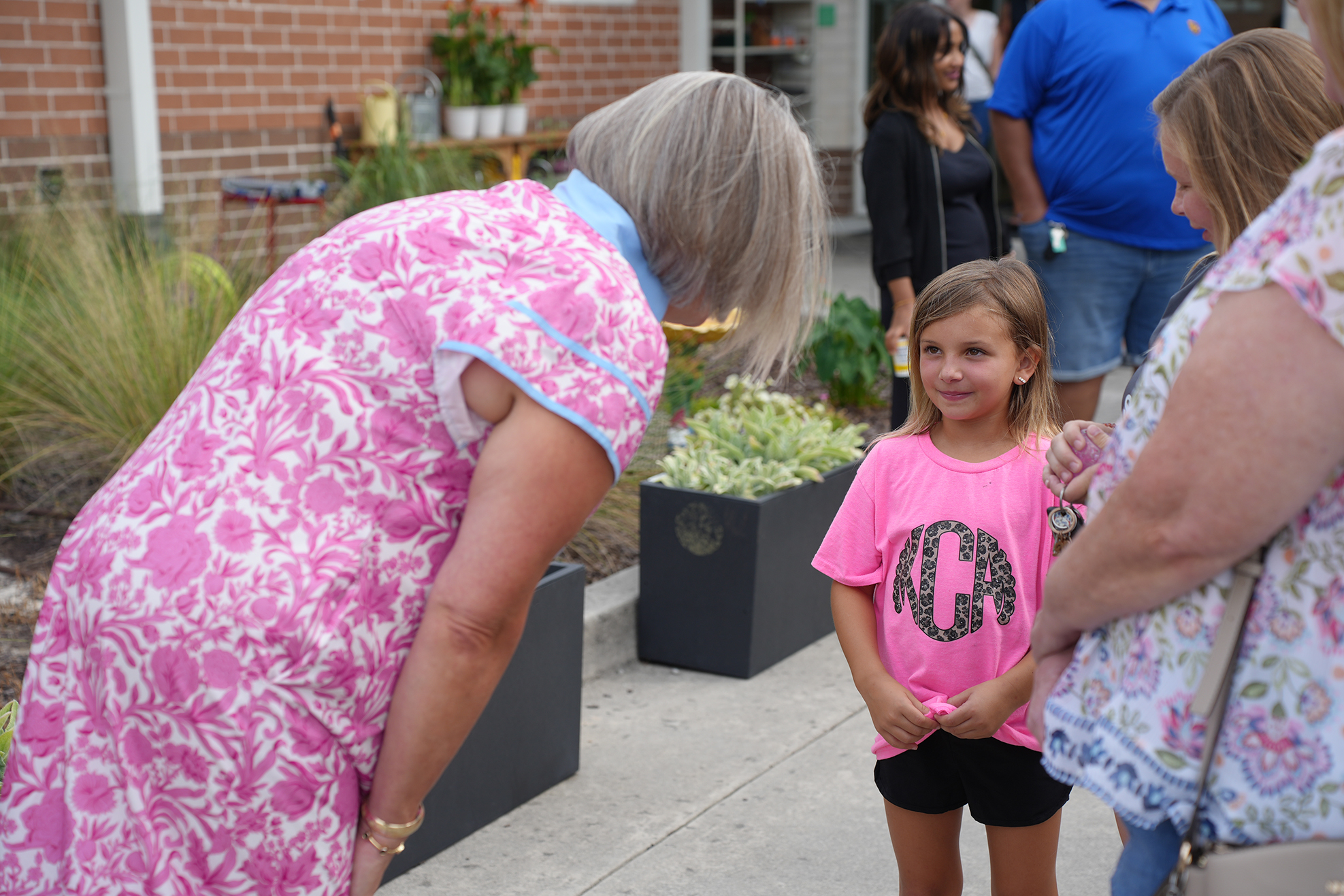 Adults and a young girl are standing in a line outside a brick building with greenery.