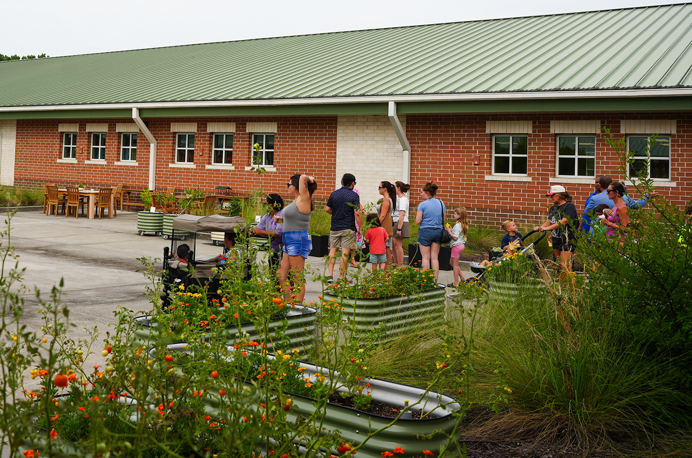 Multiple people gather outside a brick building with a green roof. A garden with various plants is in front of them.