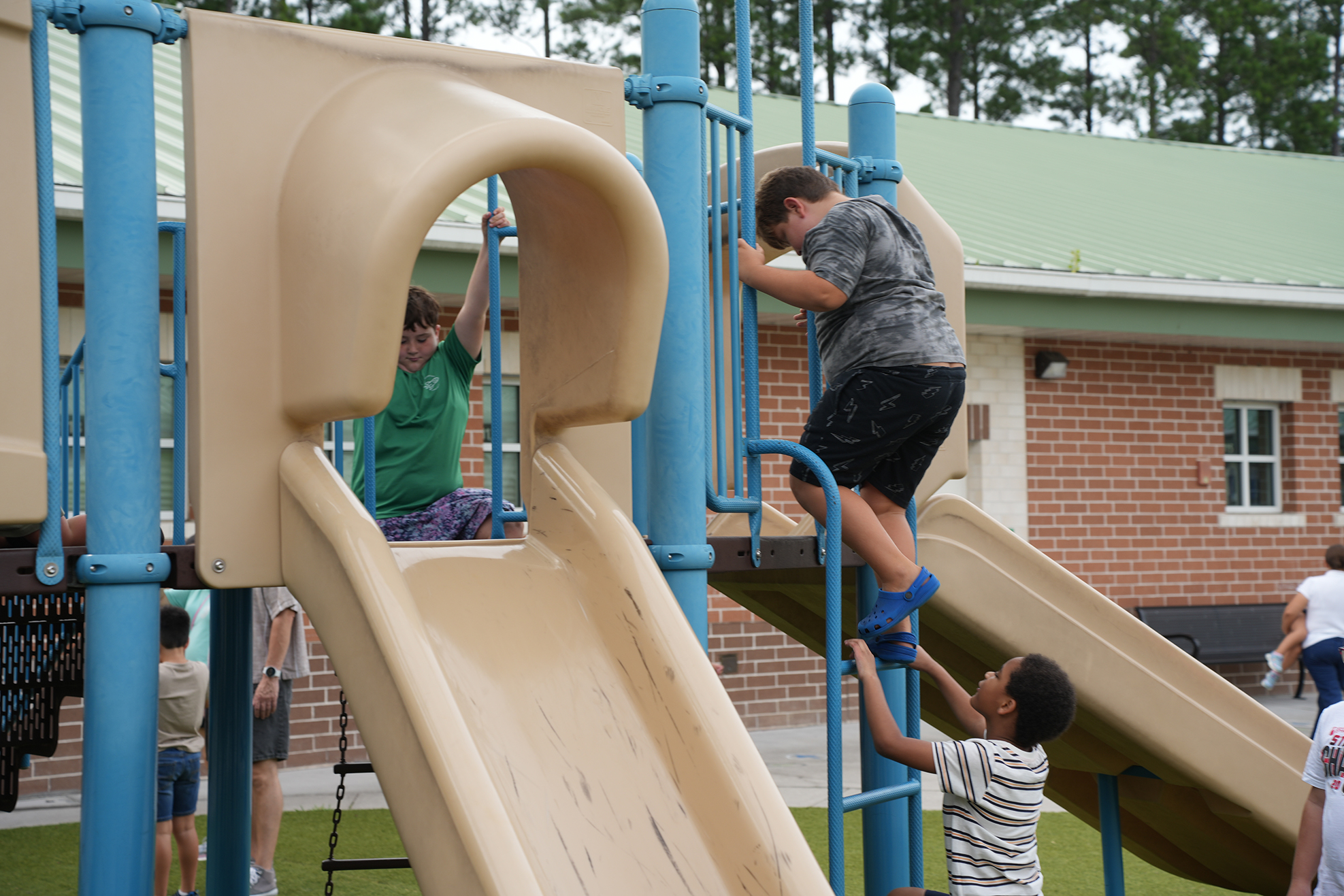 Children play on a playground with slides, one climbing up and two others on the slides. A brick building in the background.