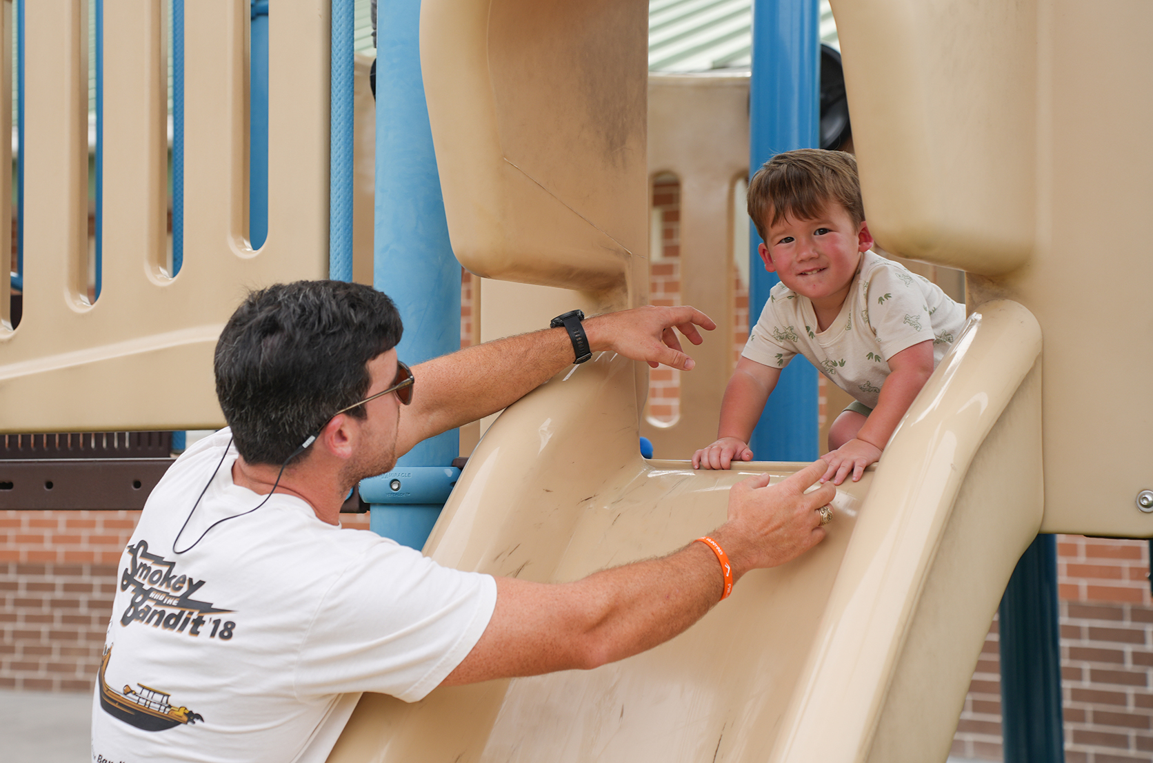 A young boy slides down a slide while a man, wearing a watch and sunglasses, helps him.