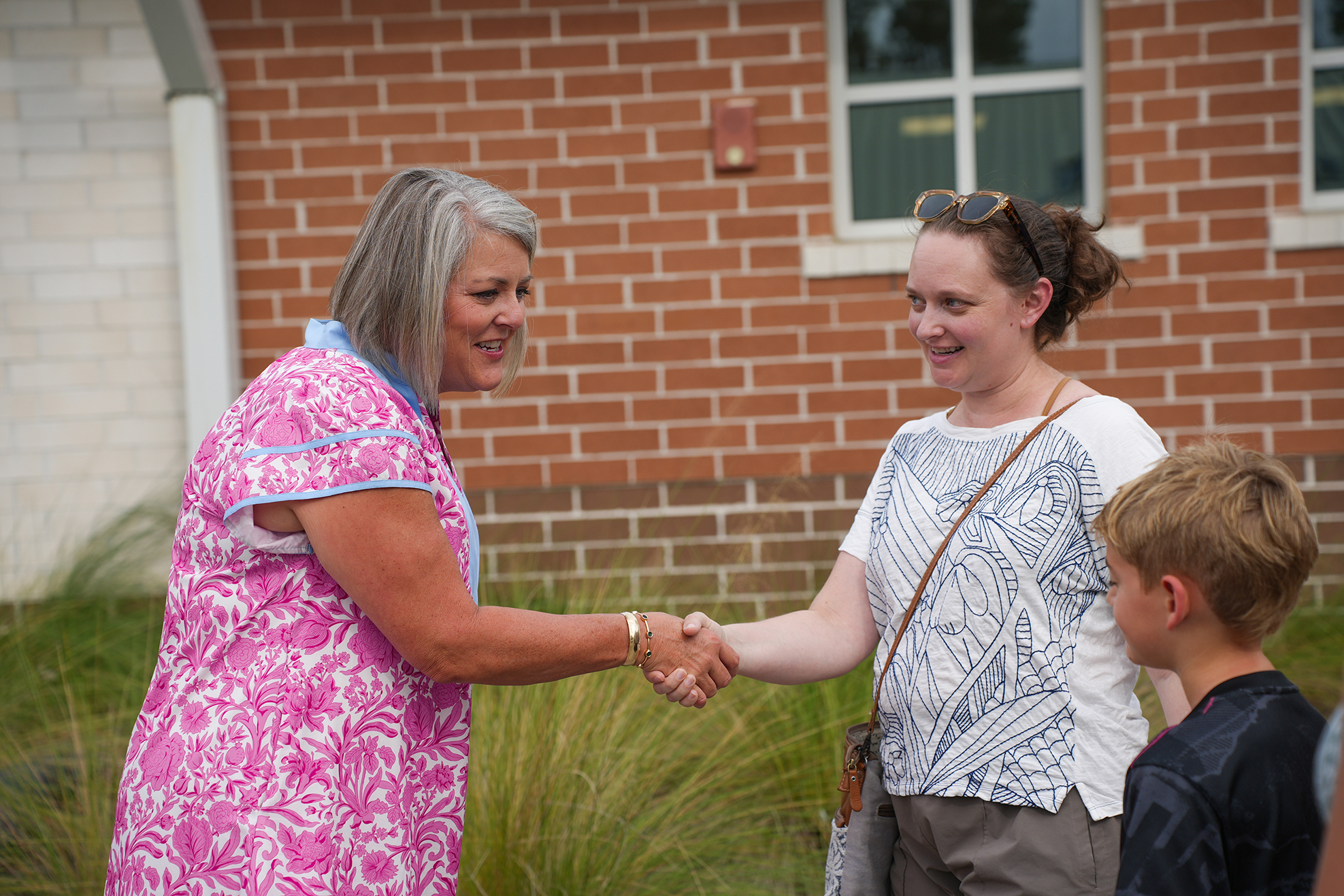 Two women shake hands in front of a brick building. One woman wears a pink floral dress. The other wears a white shirt.