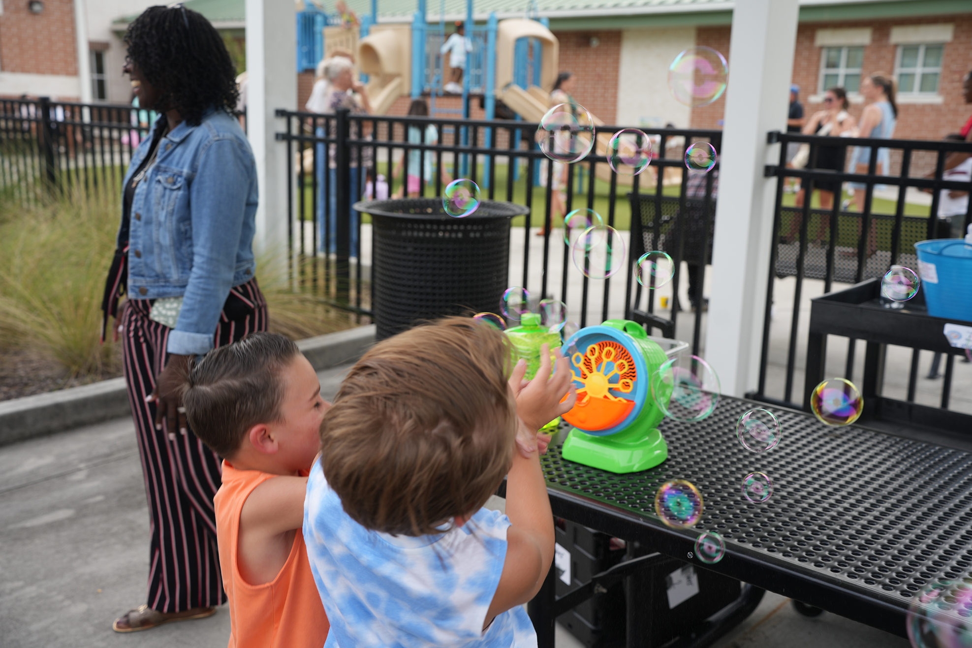A woman stands near children at a playground; they play with bubbles from a bubble machine.
