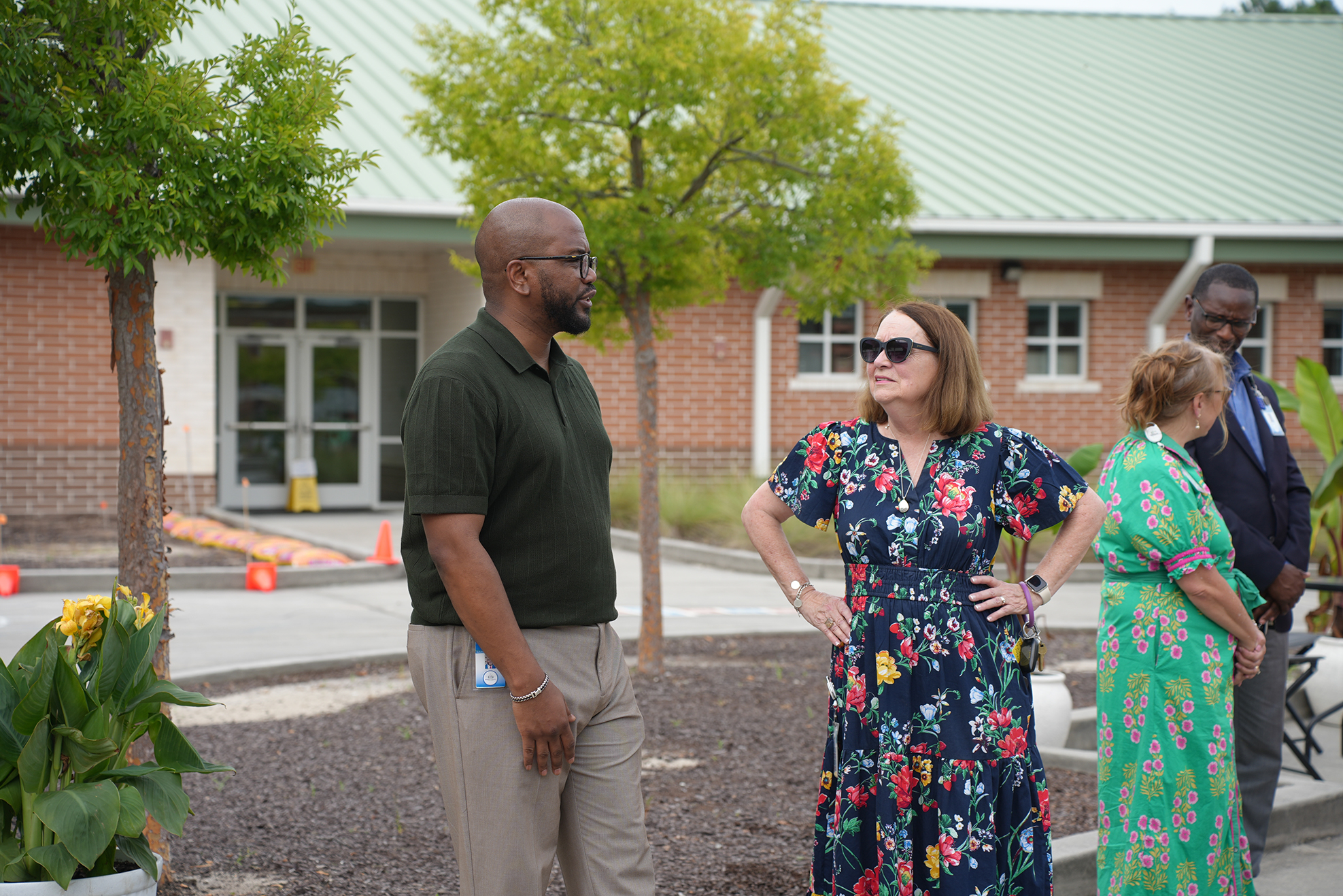 Four people in light-colored clothing stand in a park, facing a brick building with a green roof.