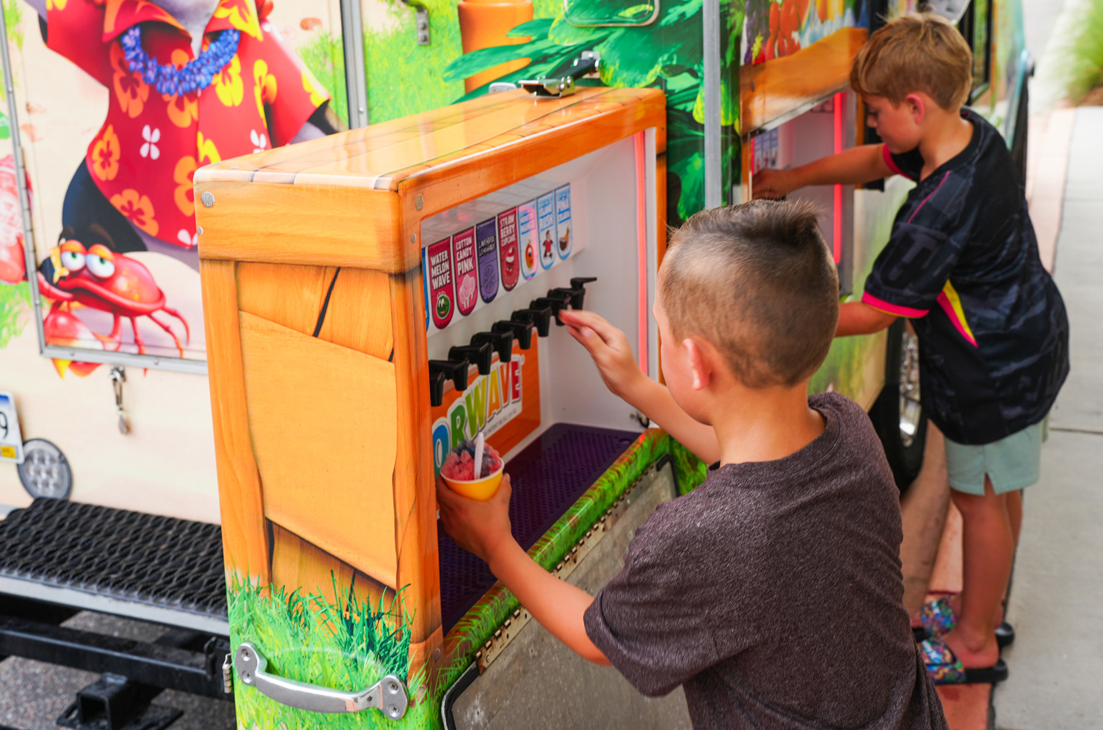 Two children use a juice machine, one holding a cup, the other selecting a drink. Brightly colored graphics are on the truck's side.