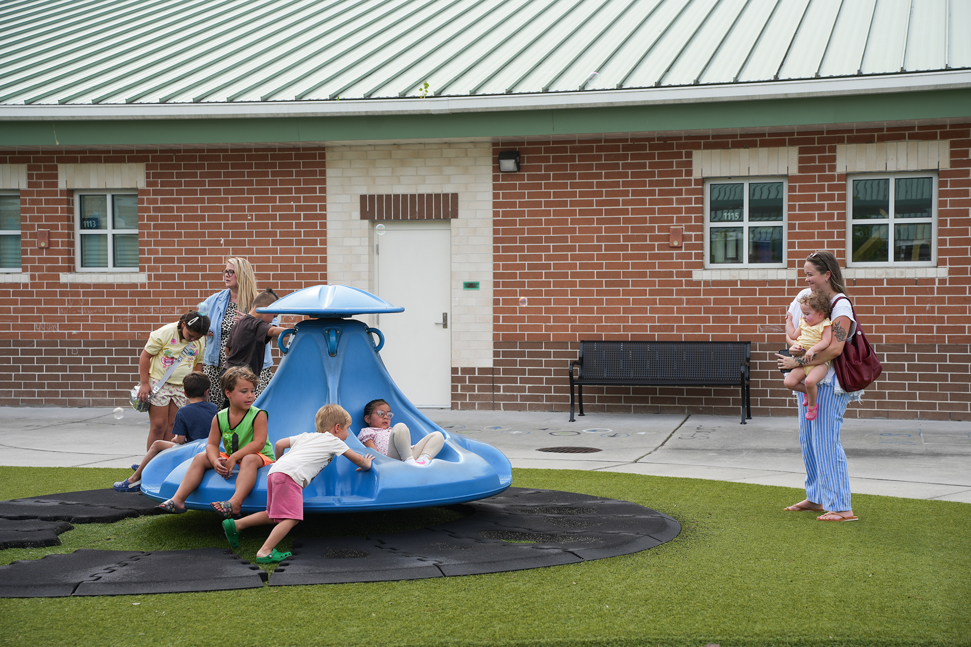 Several children play on a blue plastic spinning toy on green artificial grass. Behind them, a brick building has white trim and windows.