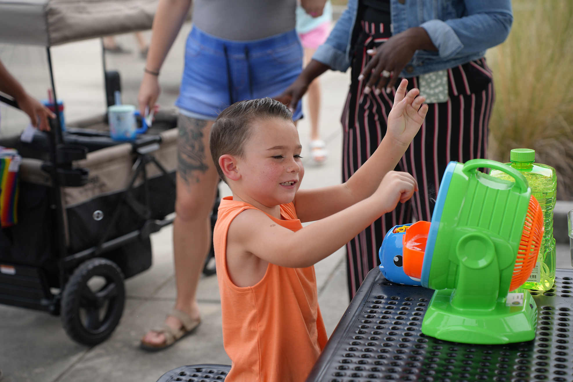 A young boy in an orange tank top reaches for a spinner on a table.
