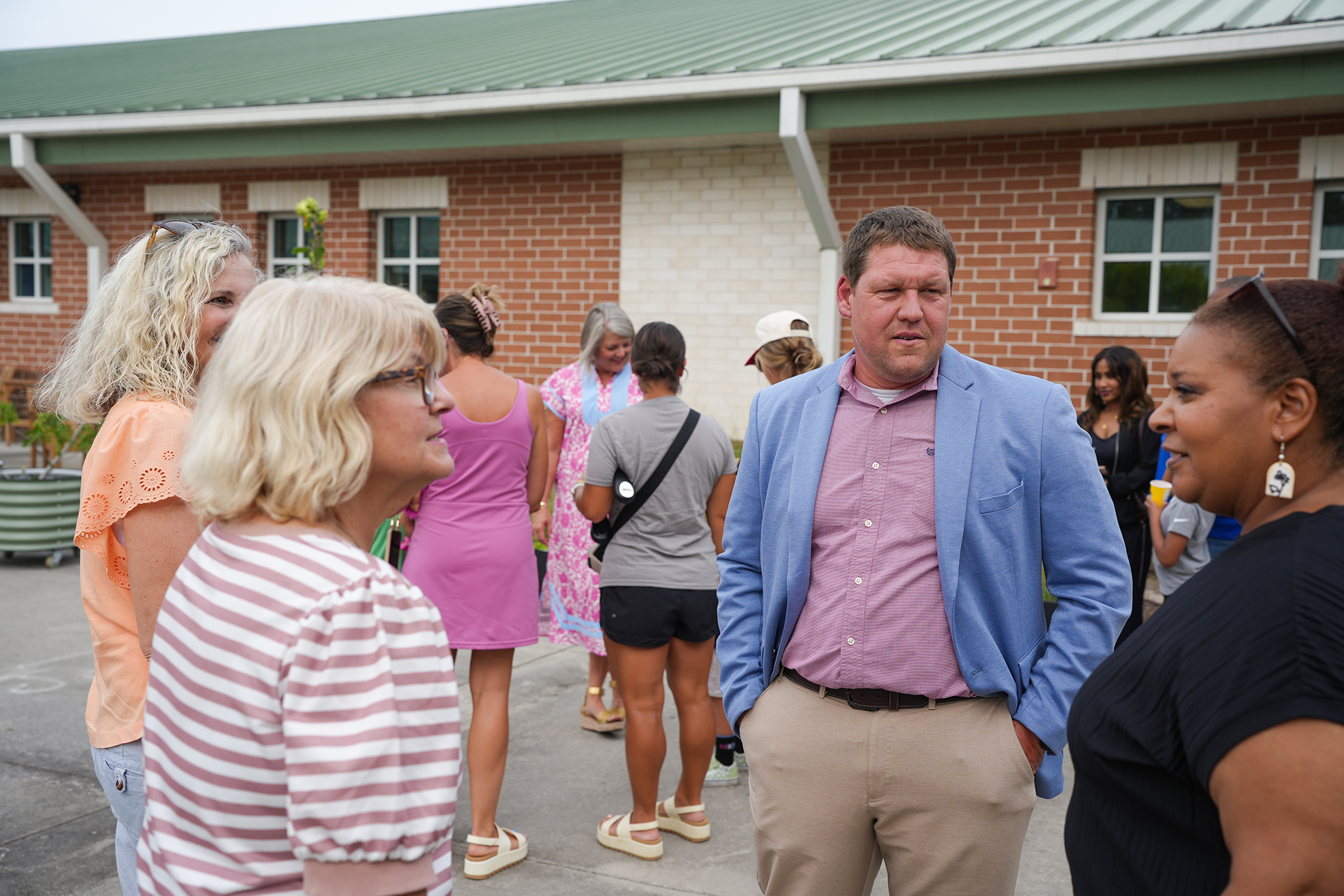 A group of adults stands outside a brick building. A man wears a blue blazer, and a woman wears a striped shirt.