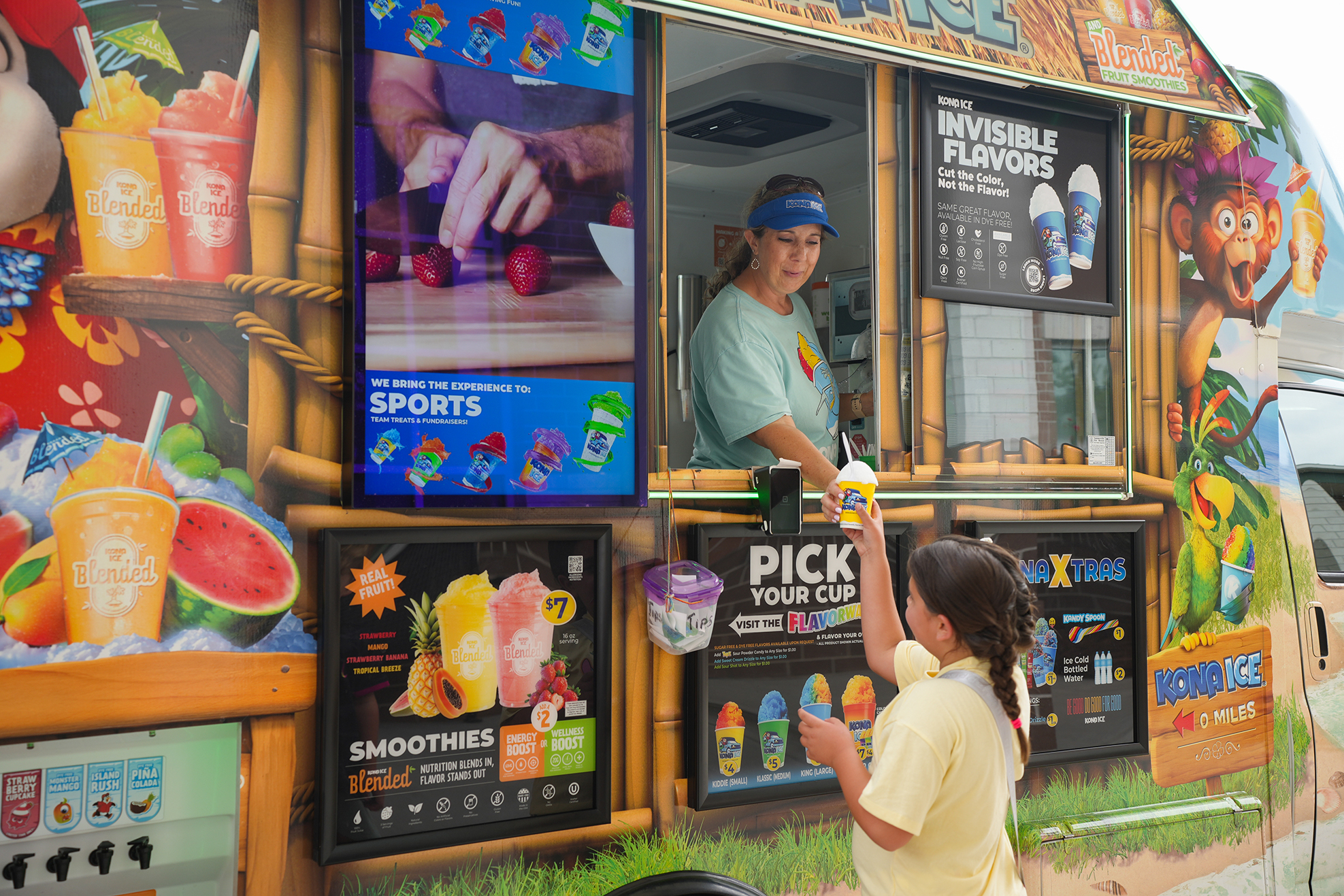 An ice cream truck with a tropical theme; a woman inside serving a young girl.