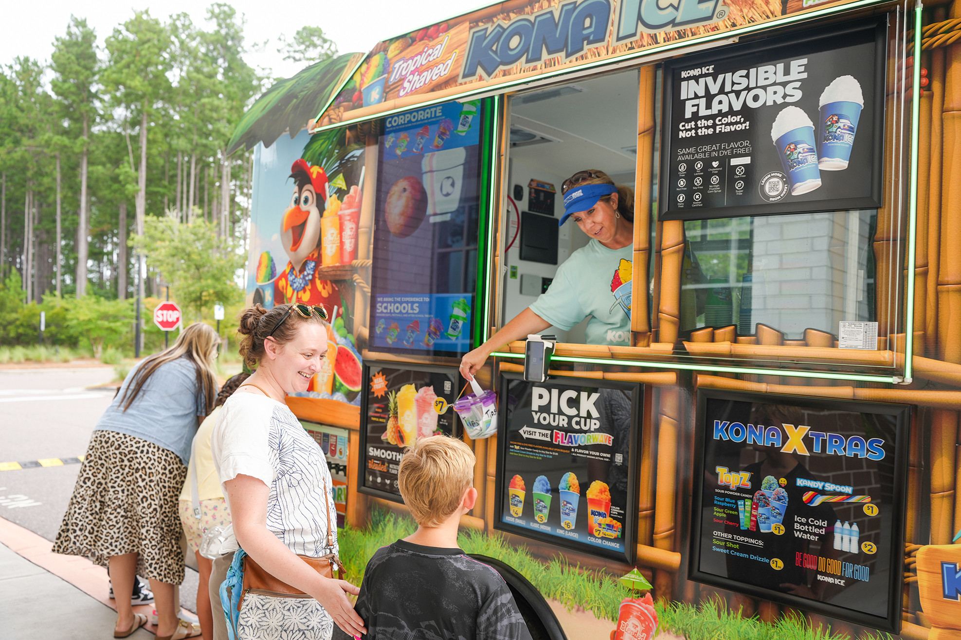 A Kona Ice truck displays various drink options. A vendor serves customers, with two adults and a child waiting. The backdrop includes trees and a street sign.
