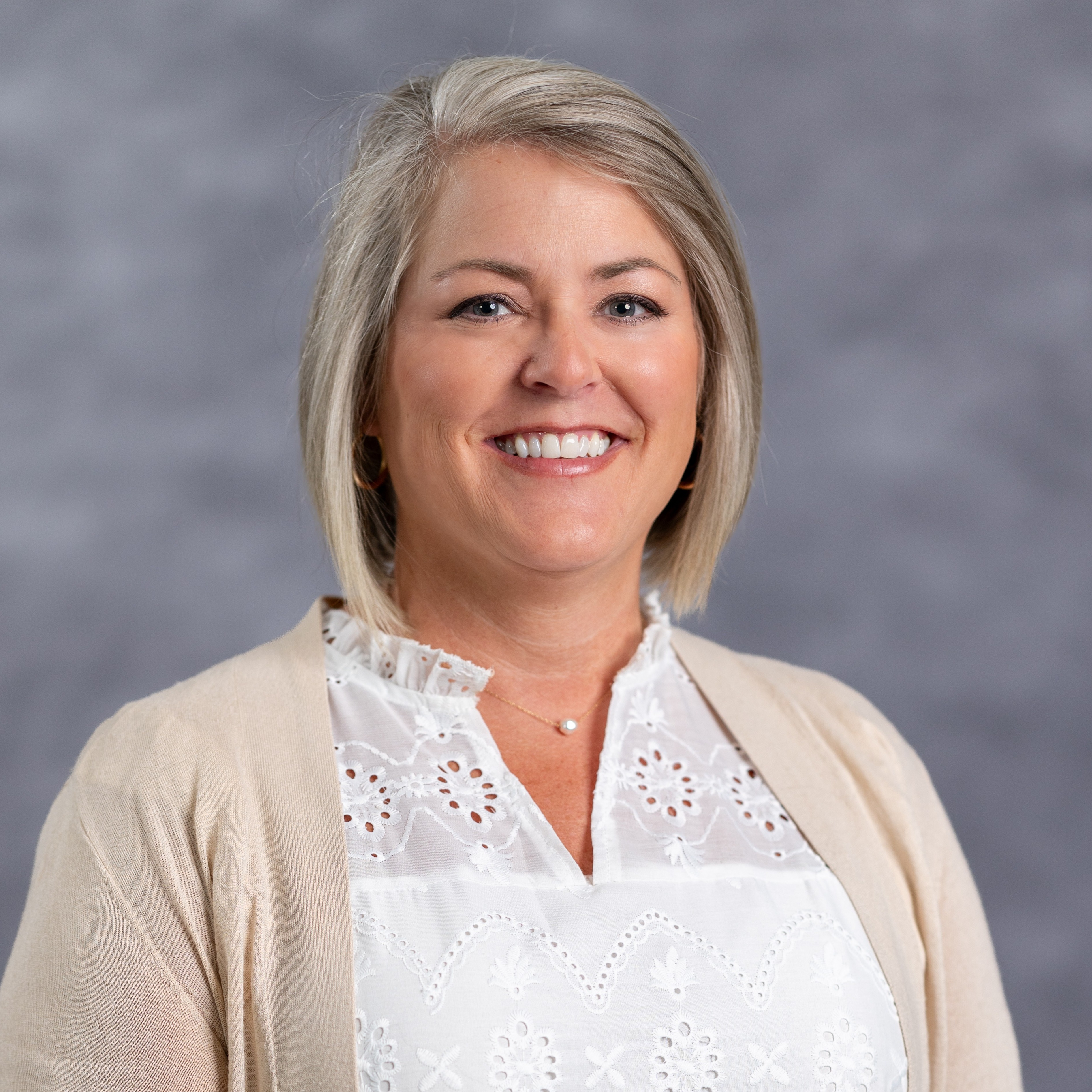 A woman with short blonde hair smiles, wearing a white blouse with lace and a beige cardigan.