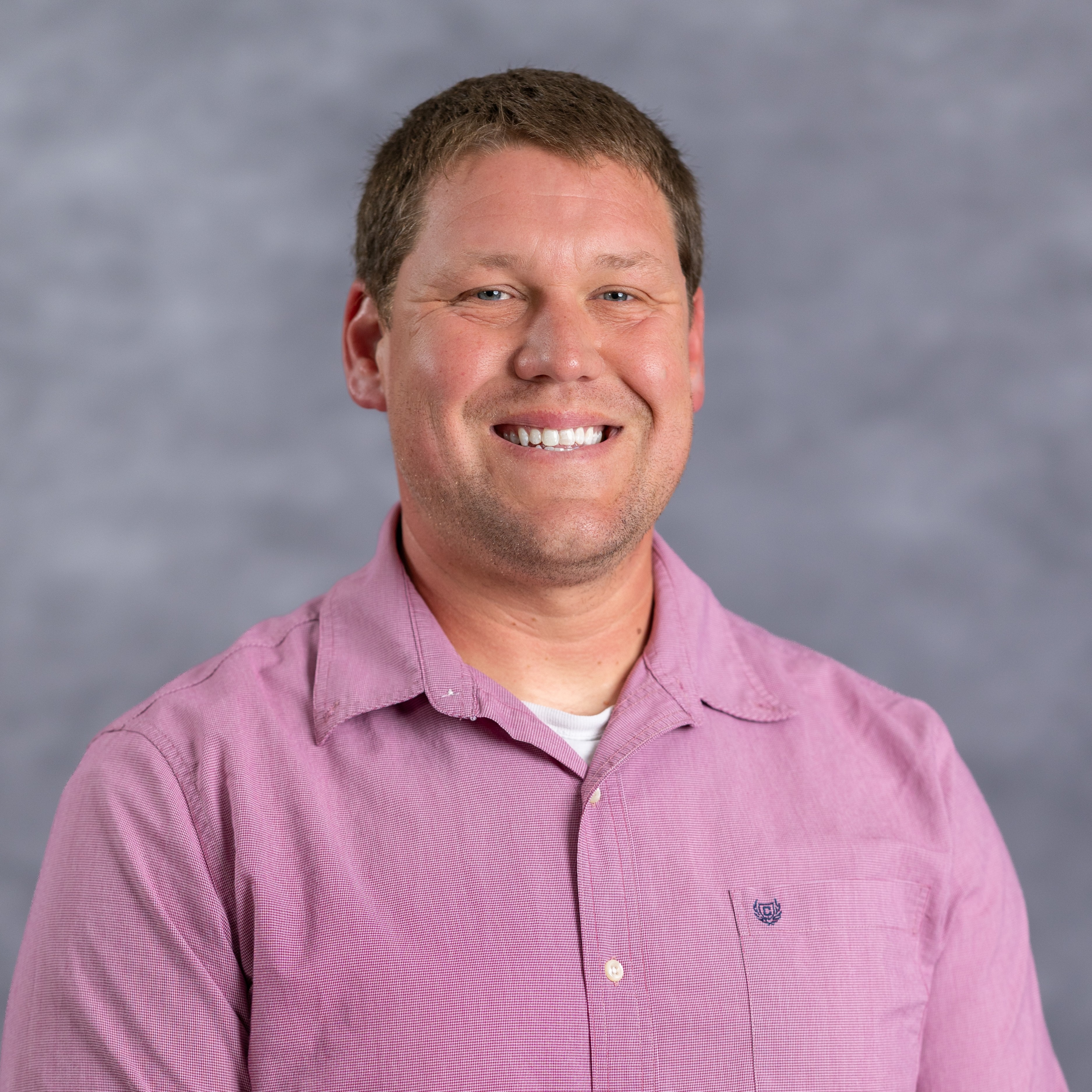 A man in a pink polo shirt with a logo on the left chest area, standing against a gray background.