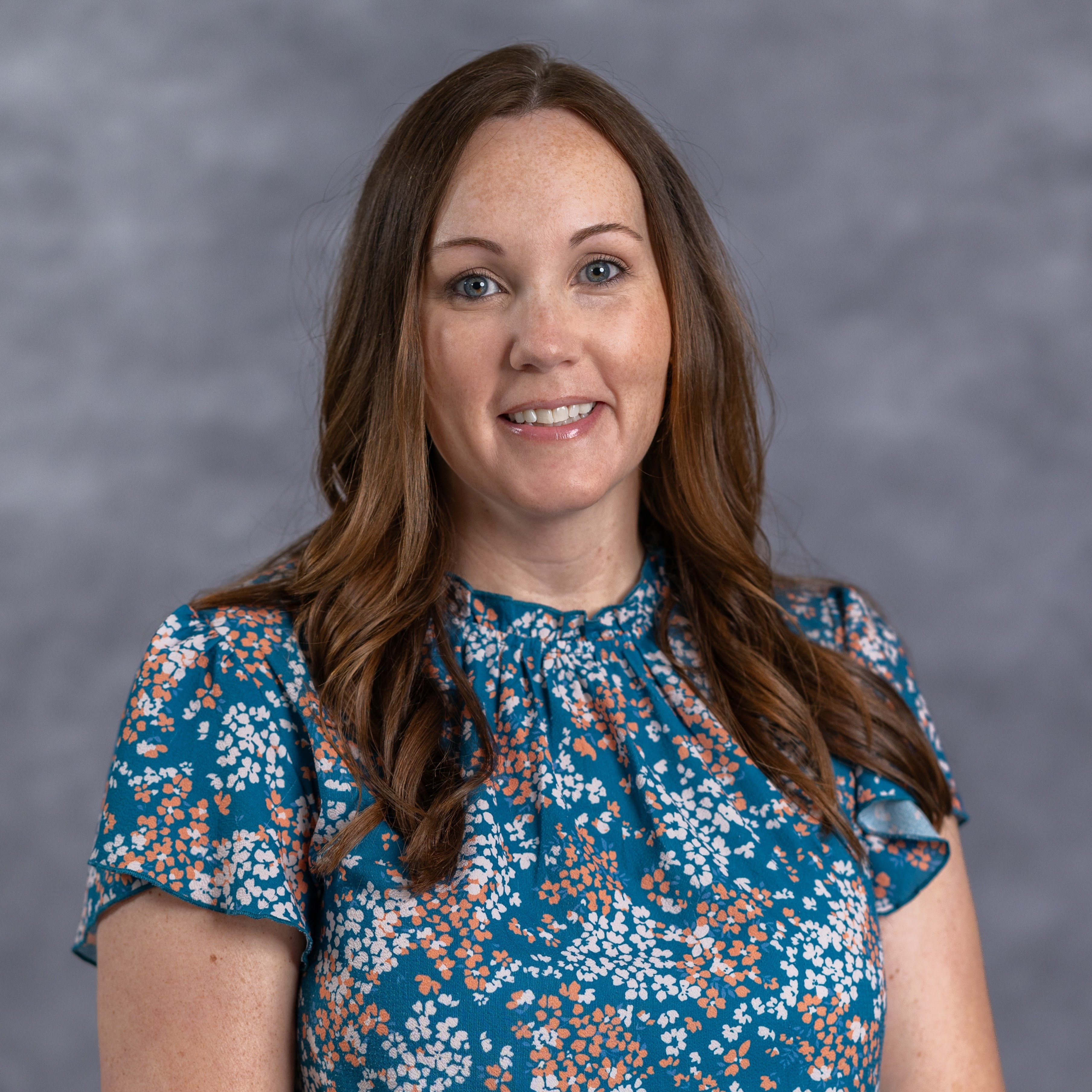 A woman with brown hair smiles, wearing a blue floral blouse, against a plain gray background.