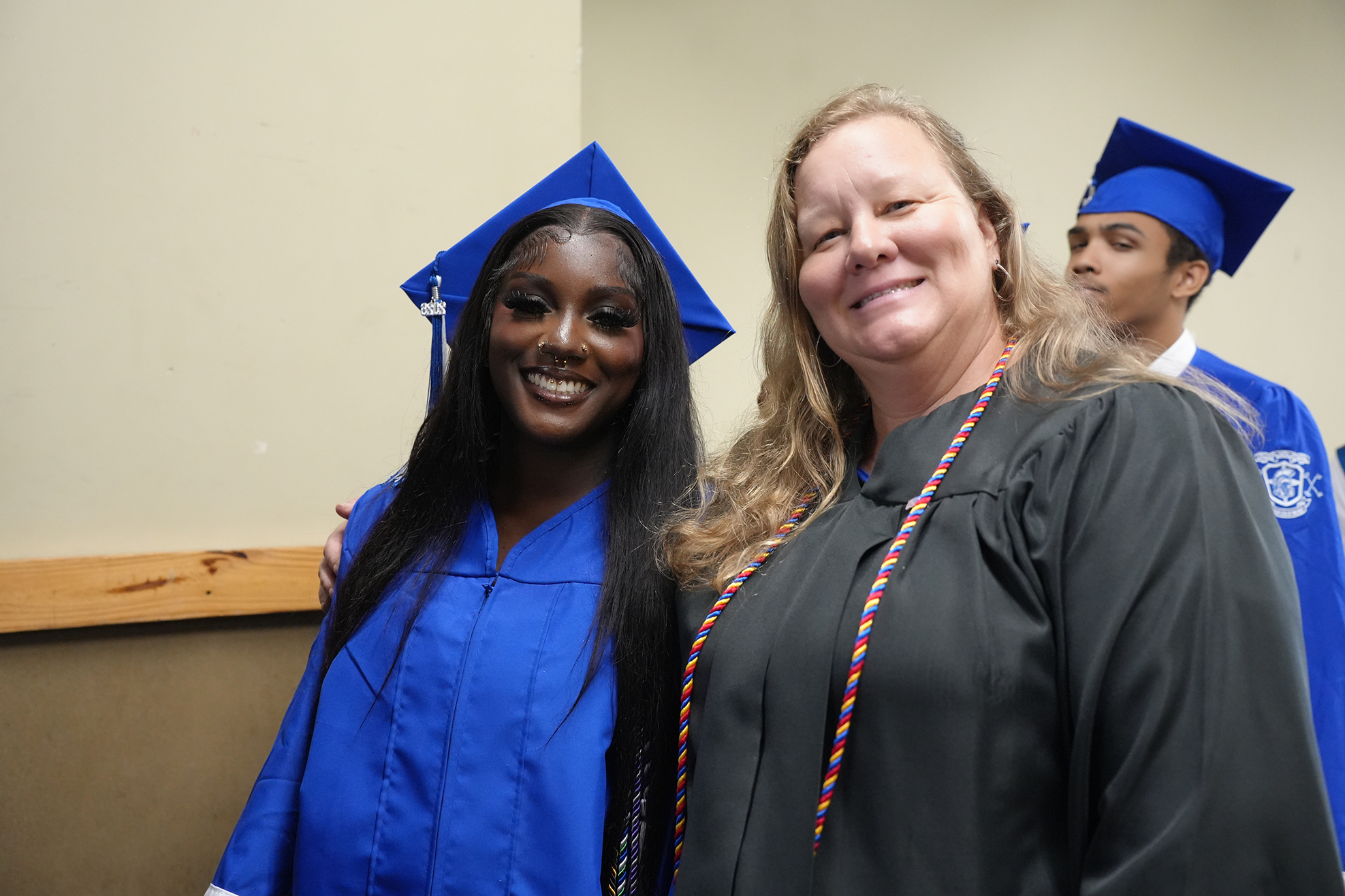 A woman in a blue gown and cap poses with a smiling woman in a black gown. Another individual in a blue cap and gown stands behind them.