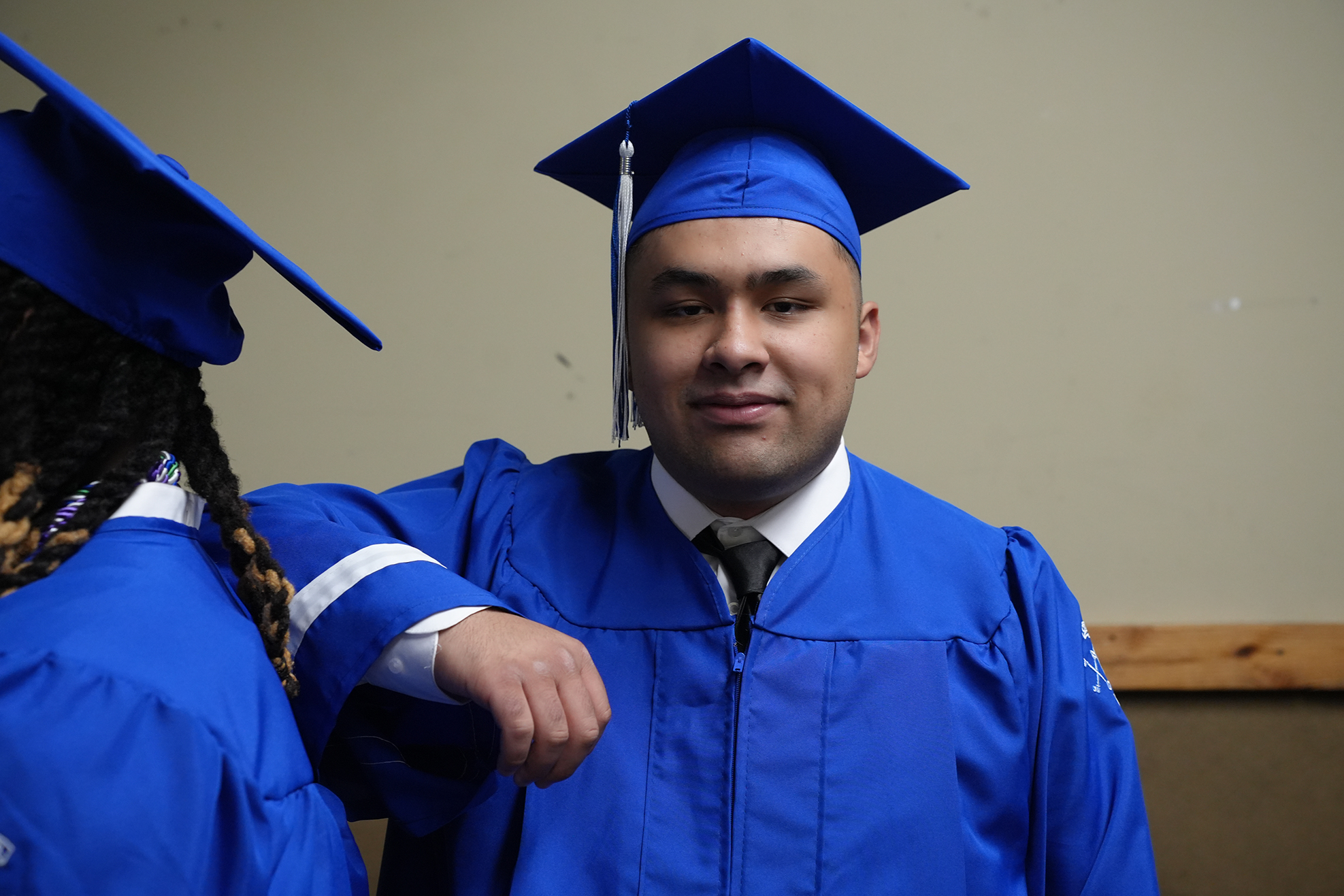 Two people in blue graduation gowns and caps stand close. One with hand on shoulder of the other.