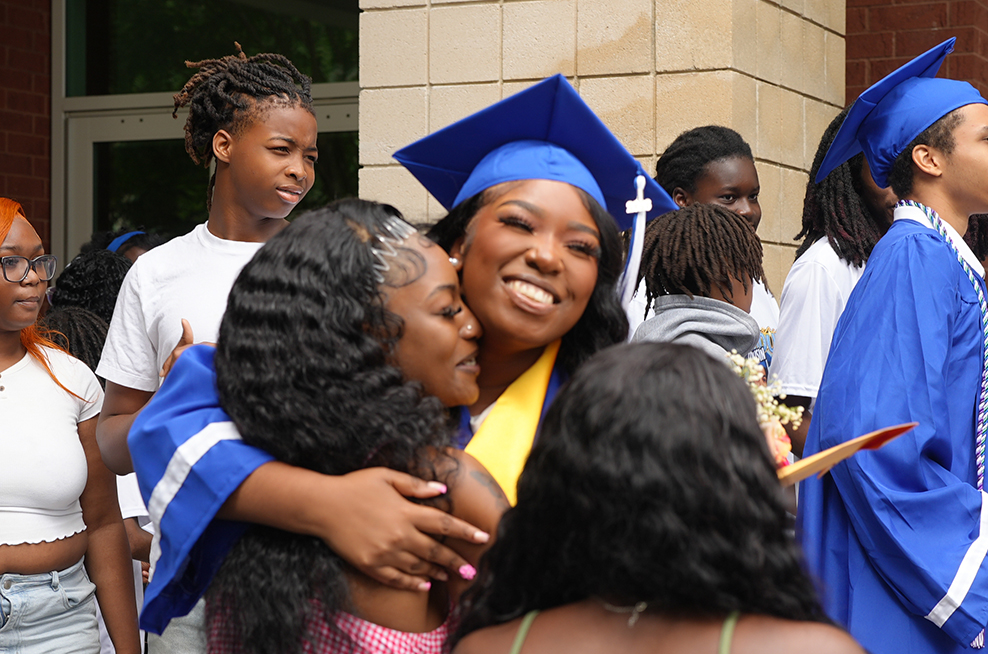 A group of people, some in graduation gowns, are gathered outside a building. A woman in a blue gown smiles while hugging another person.