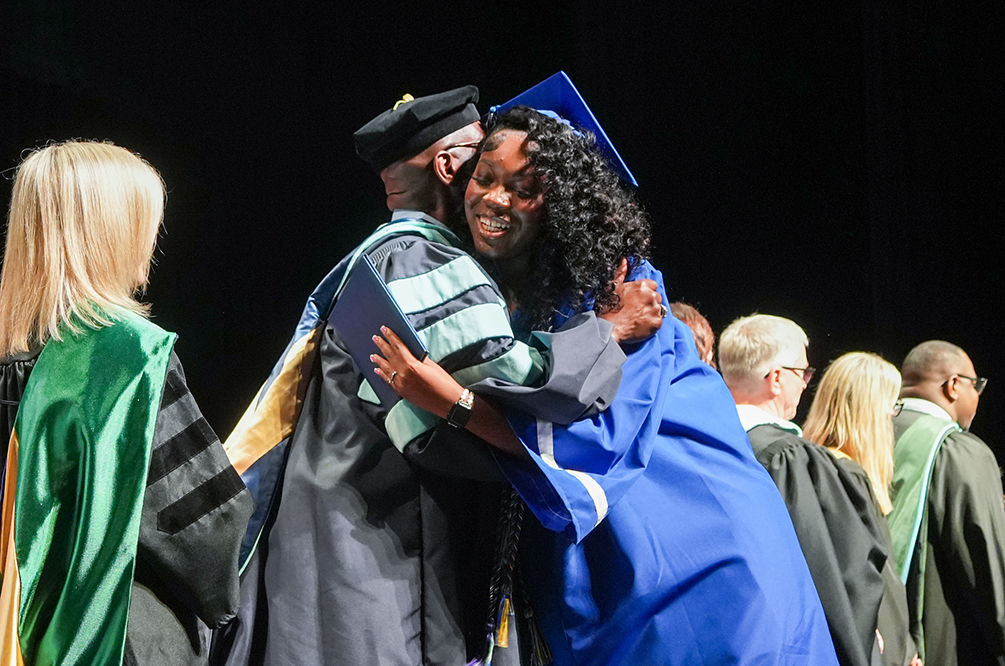 A person in a blue gown is being hugged by another in a black and gold gown. Behind them, more people in green and black gowns are standing.