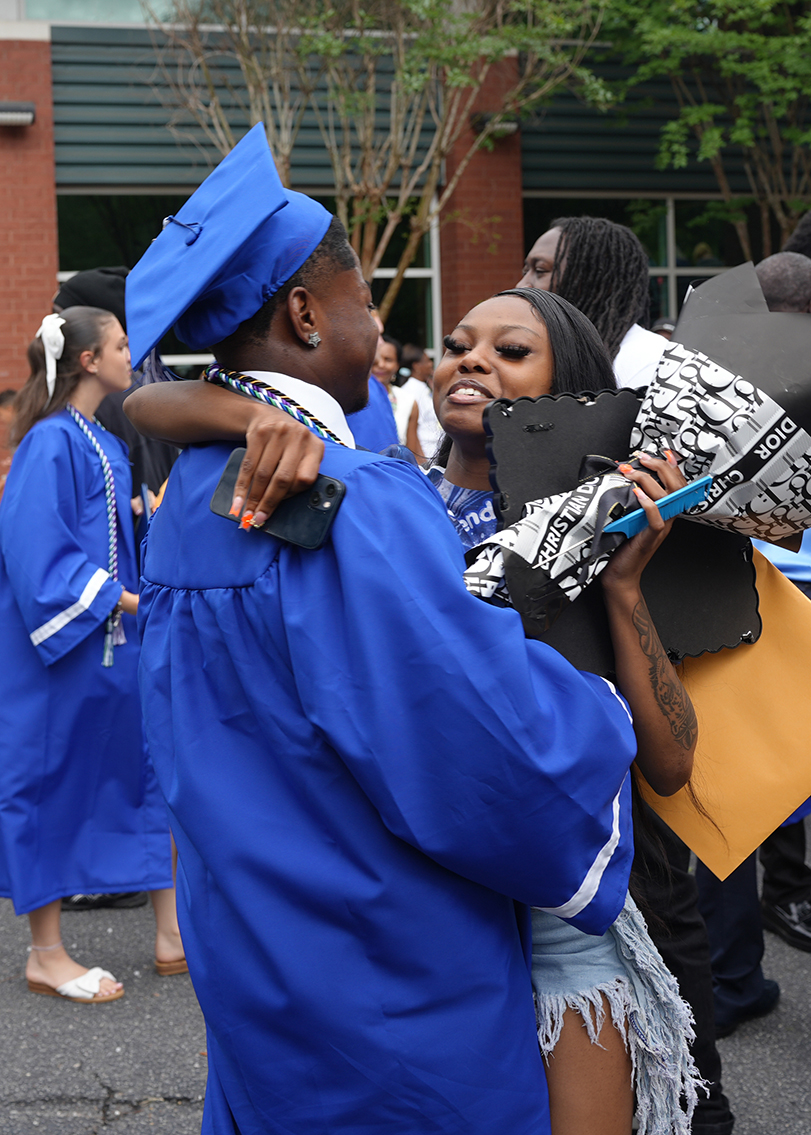 A man in a blue gown hugs a woman with a cap and gown, holding a cap. They smile.