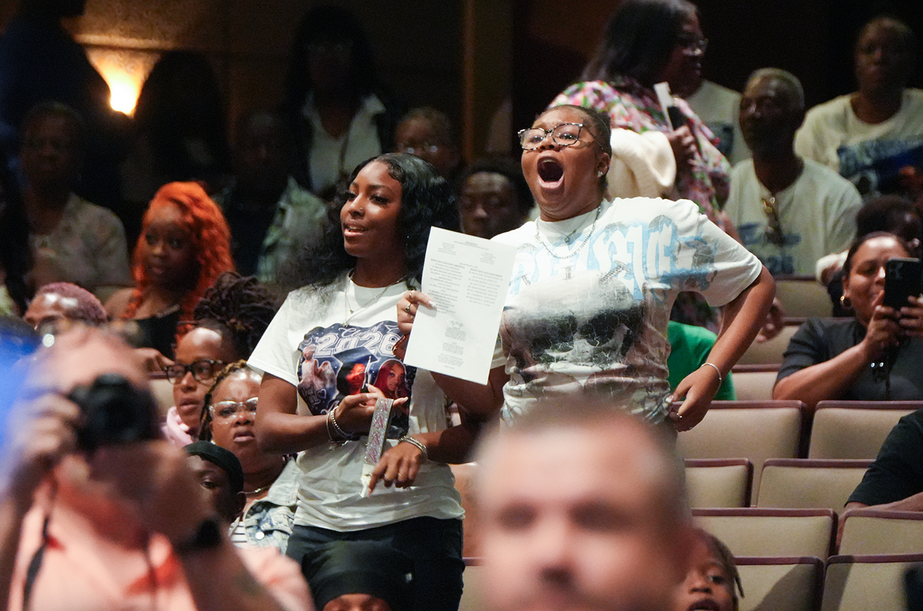 A crowd of people in an auditorium, with two women holding papers and standing, some seated.
