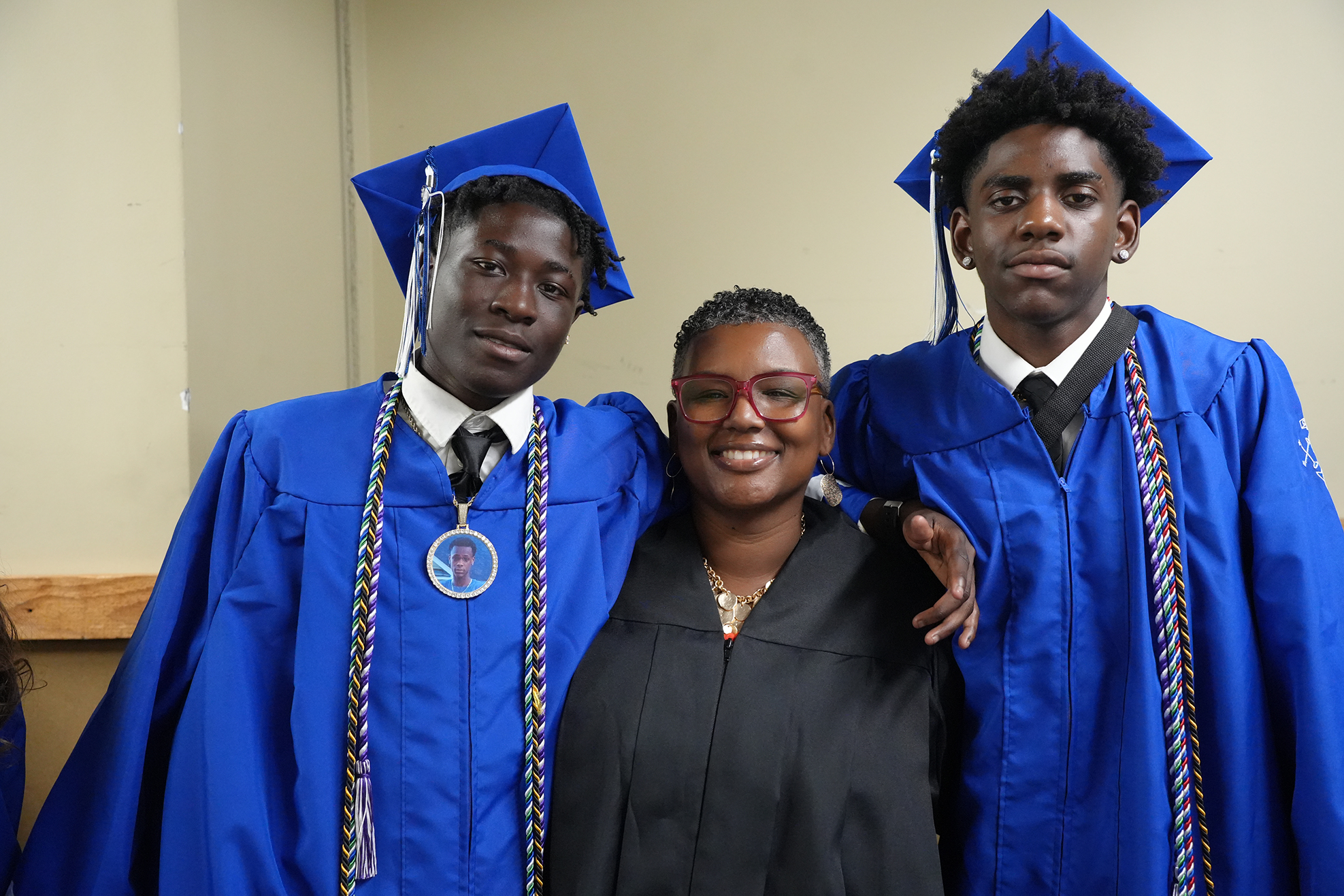 Two men in blue gowns and mortarboards with a woman in a black gown, smiling, standing in a room.