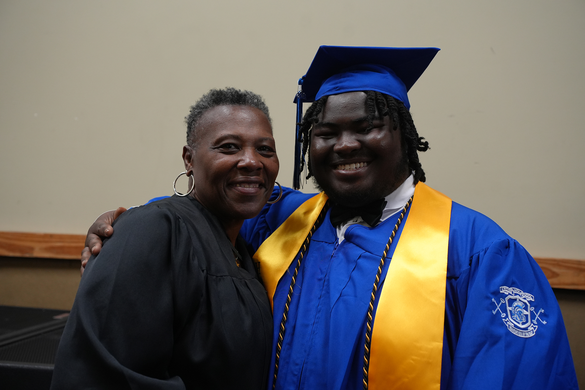 A smiling adult man in a blue graduation gown stands next to a woman in a black robe.