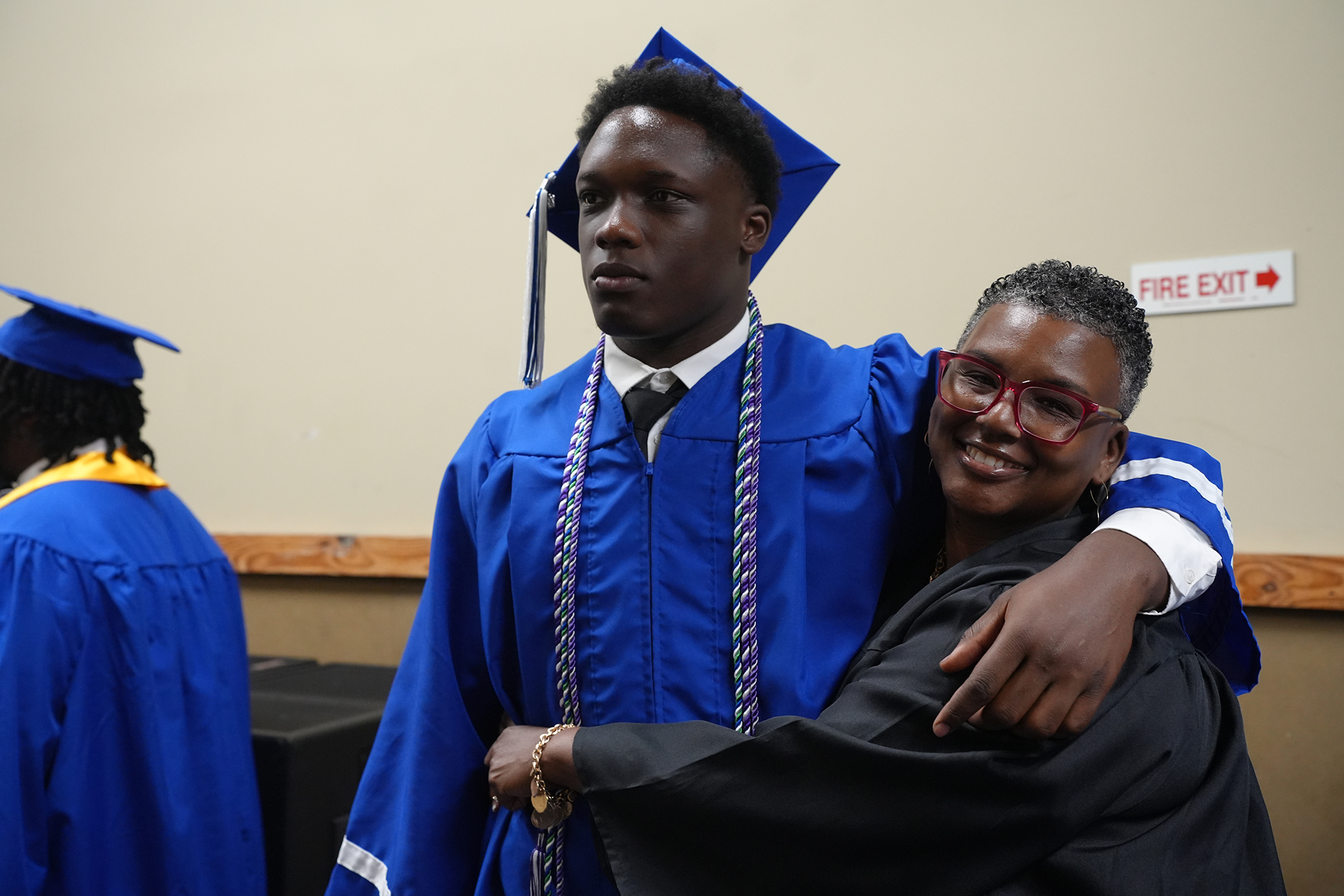 A man in a blue graduation gown and cap embraces a woman in a black gown and cap.