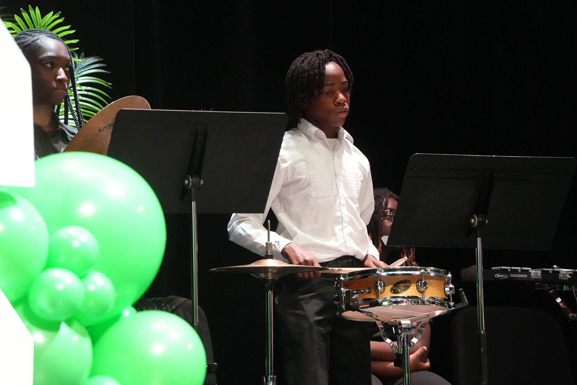 A young boy playing drums on stage with green balloons in the foreground and two people seated behind him.