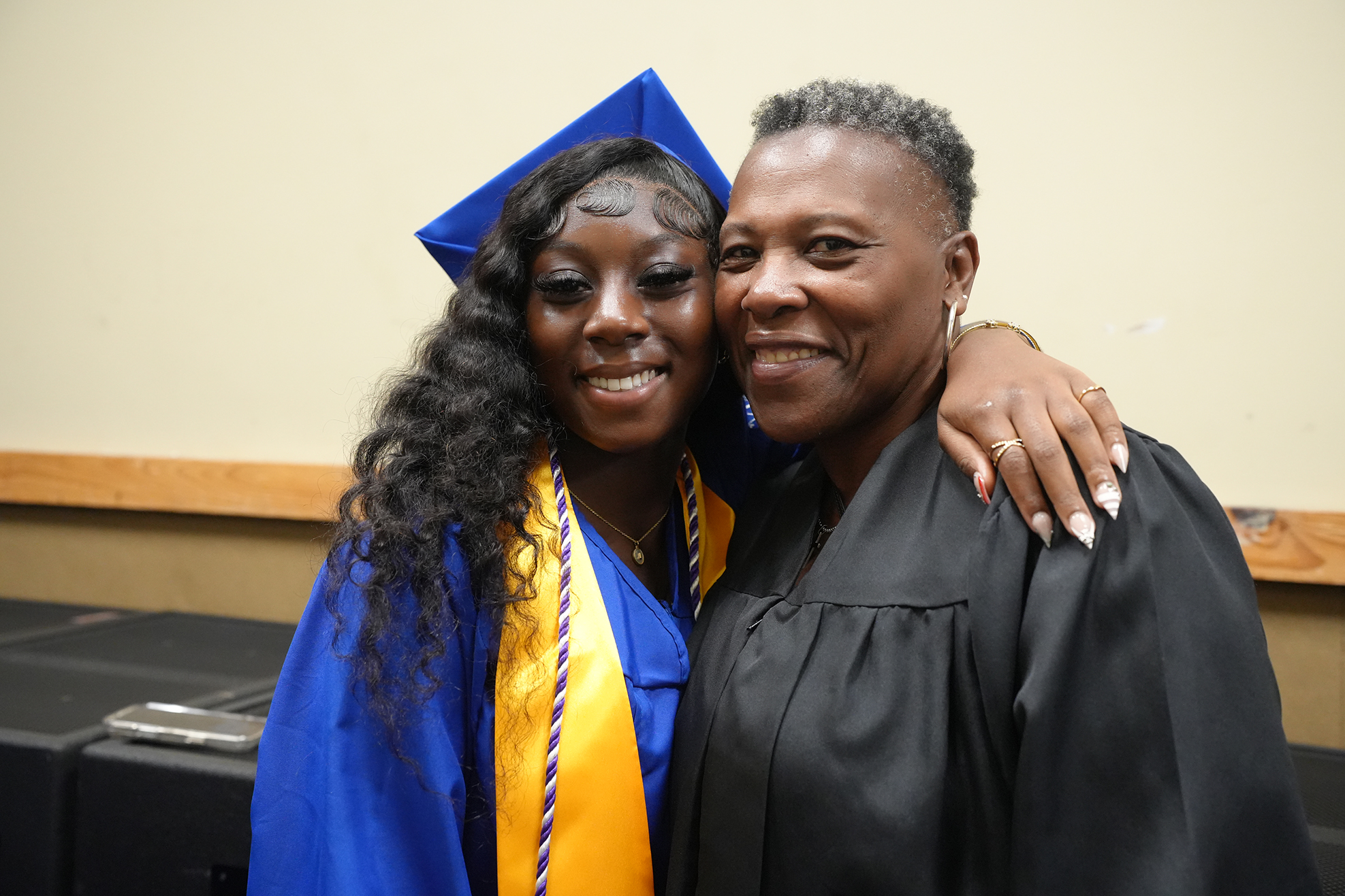 Woman in a blue cap and gown posing with a woman in a black gown, both smiling.