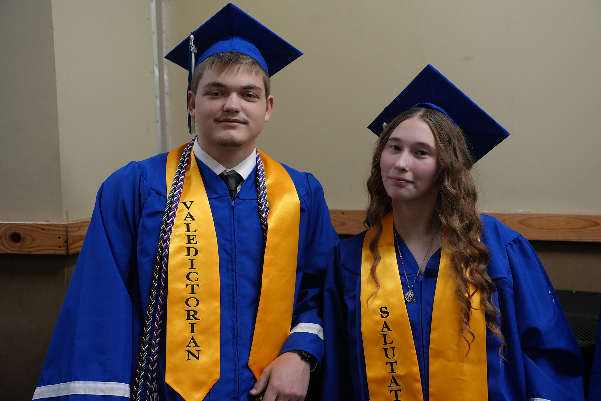 Two individuals in blue and yellow graduation attire, one with a sash reading "Valedictorian" and the other with "Salutatorian."